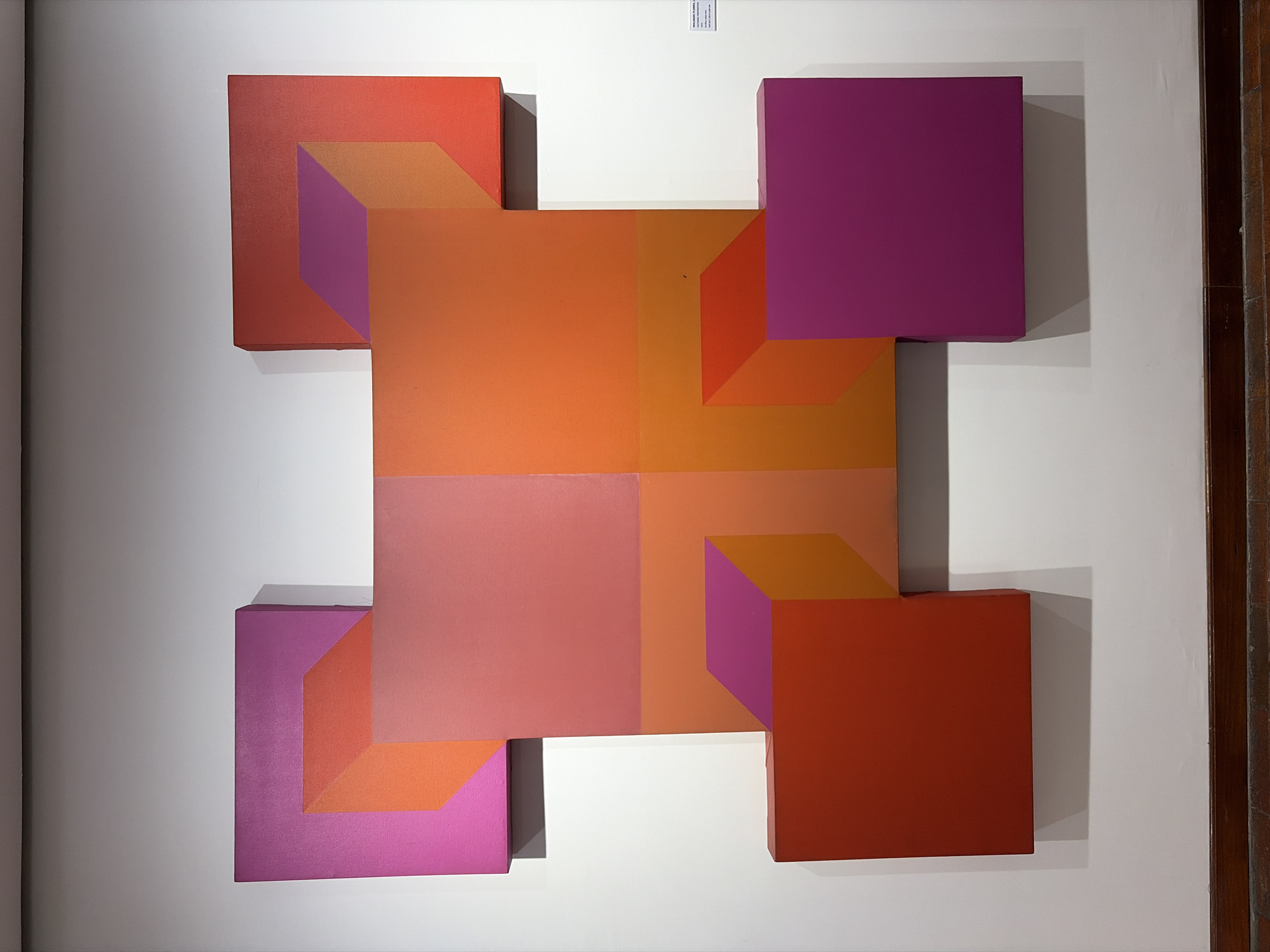 A photograph shows a vibrant abstract geometric artwork mounted on a white wall. The artwork comprises a central square divided into four smaller squares with an orange to peach gradient. Attached to each side of the central square are smaller projecting squares in red and magenta hues, creating a three-dimensional illusion. The artwork casts shadows on the wall, enhancing its perceived depth. A small white label with black text is affixed to the wall above the artwork. A dark brown door frame is visible on the right side of the image. The precise location within Ciudad Bolivar, Venezuela is not discernible from the image. No people or subjects are present in the photograph.