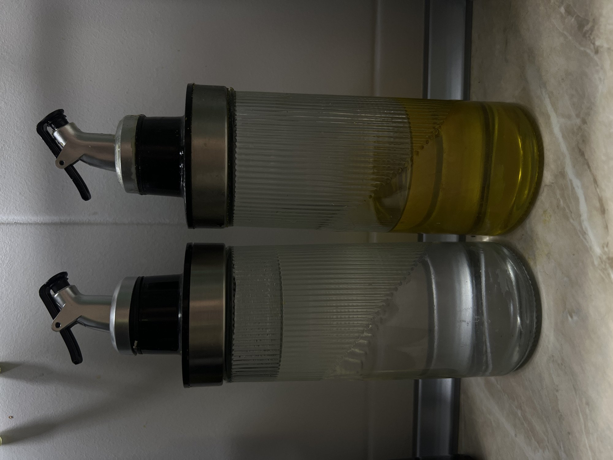 Two glass bottles with spouted tops stand on a speckled countertop. The bottle on the left contains a clear liquid, likely water or vinegar, filled to about two-thirds of its capacity. The bottle on the right contains a yellow liquid, presumed to be cooking oil, also filled to approximately two-thirds of its capacity. Both bottles have ribbed glass exteriors and are capped with black plastic collars topped by metallic pouring spouts with black levers. The background consists of a tiled wall and a dark horizontal element, possibly a backsplash or shelf.