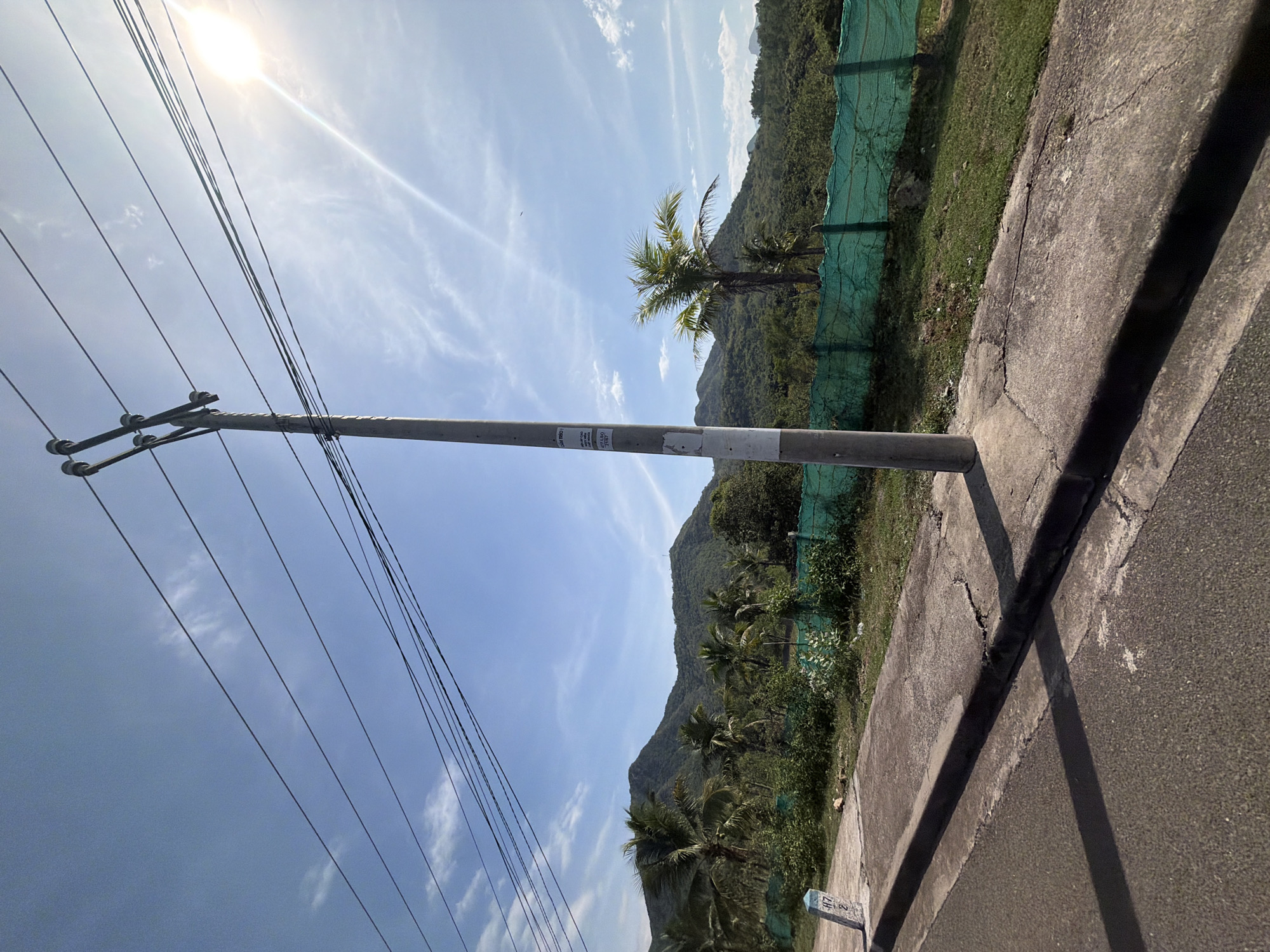 The image captures a roadside scene in Vĩnh Lương, Vietnam, under a bright, partly cloudy sky. A concrete utility pole, adorned with multiple wires and insulators, stands prominently in the foreground, casting a shadow on the adjacent pavement. To the left, a paved road runs alongside a raised concrete sidewalk with a curb. A small, blue and white marker post with the inscription "H17 2" is visible on the sidewalk.
Beyond the sidewalk, a stretch of green grass and vegetation is bordered by a vibrant green mesh fence. In the background, lush, tree-covered mountains form a picturesque backdrop. Several palm trees, characteristic of the tropical climate, are scattered throughout the landscape, adding to the scenic beauty. The sun is high in the sky, with its rays creating a lens flare effect, indicating a clear and sunny day. The overall impression is one of a rural or semi-rural area, possibly on the outskirts of Vĩnh Lương, with infrastructure for electricity and a natural mountainous environment.