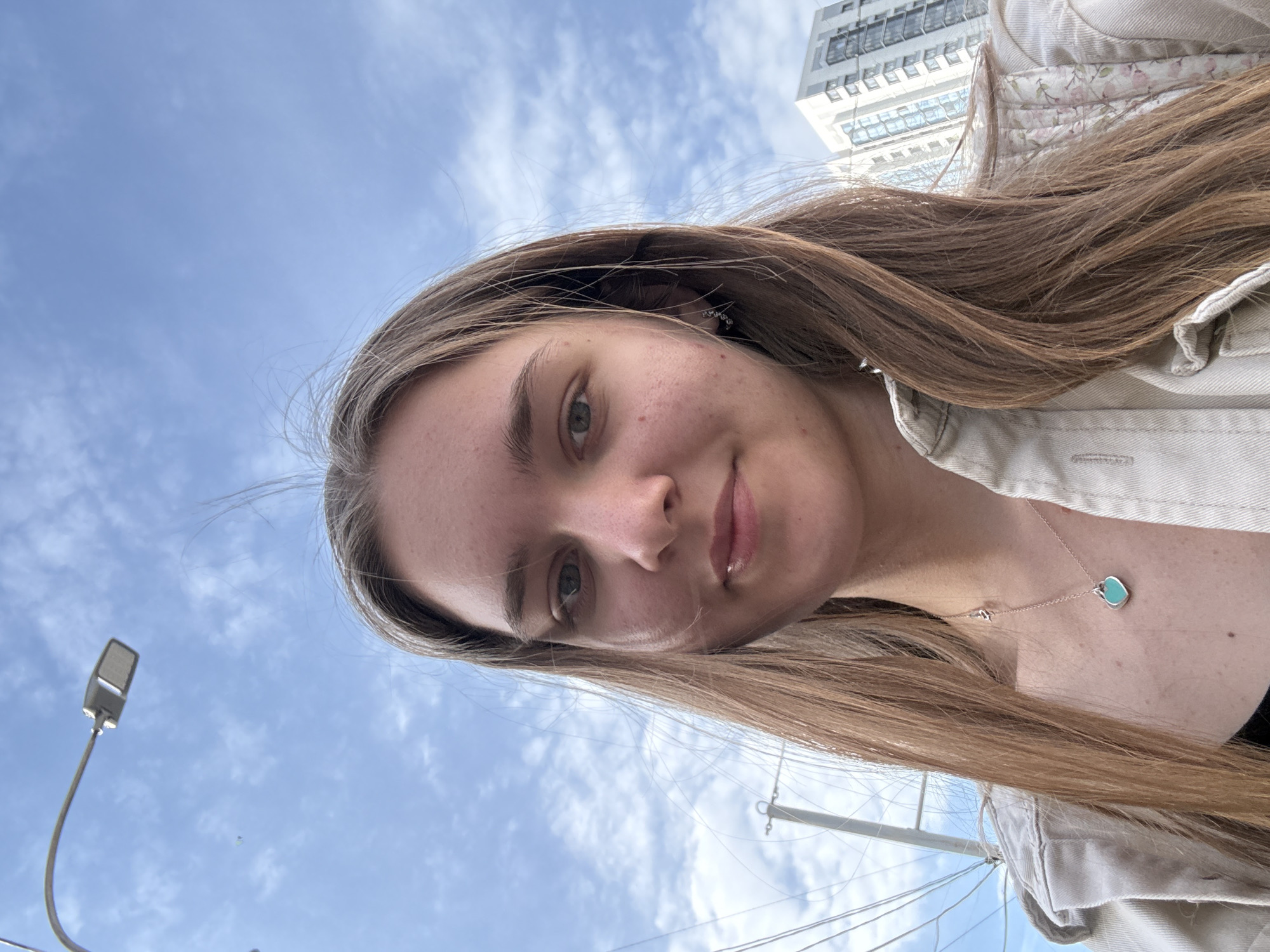 A low-angle, medium close-up selfie of a young woman with light brown hair, fair skin, and light-colored eyes. She is wearing a light-colored jacket and a necklace with a heart-shaped pendant. A street lamp is visible on the left side of the frame. In the background, there is a light blue sky with patchy clouds. In the distance, the upper portion of a tall, modern building can be seen, with rows of windows. Based on the architectural style and environment, it is likely that the photo was taken in Perm, Russia. Electrical lines and poles are also visible in the background.