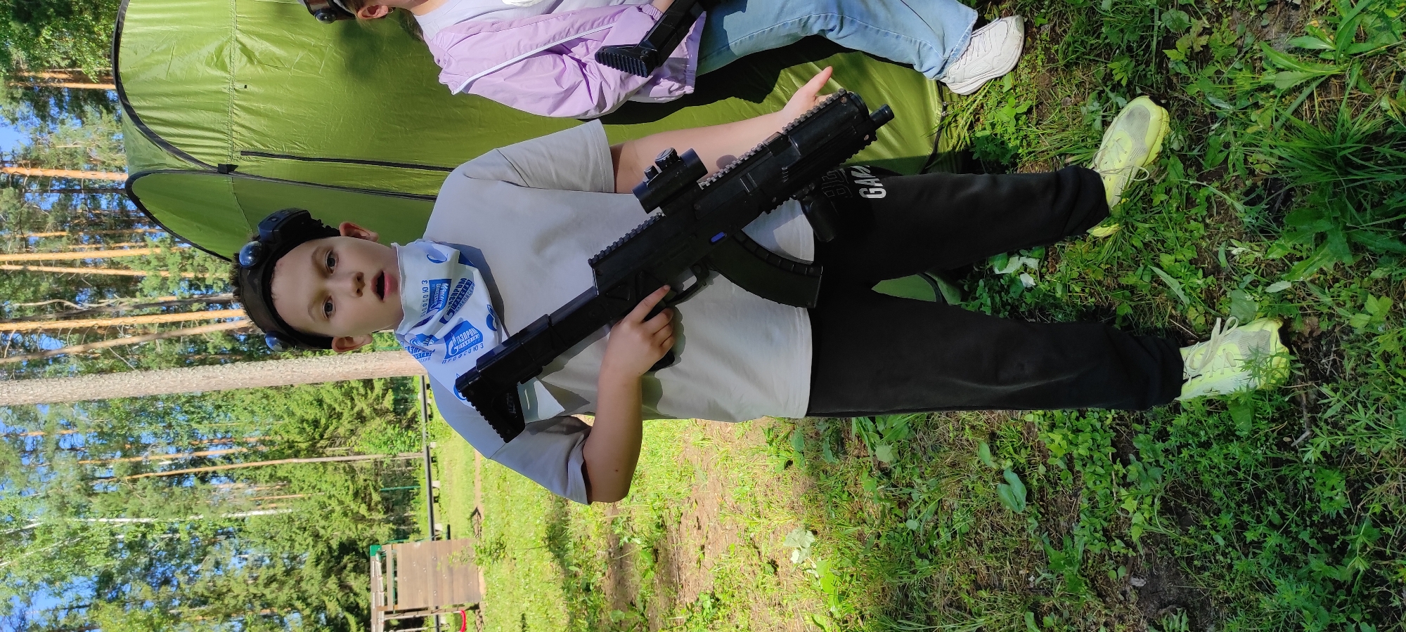A young boy, wearing a grey t-shirt, black pants, and bright yellow sneakers, stands in a grassy area outdoors, holding a black toy rifle. He has a black headset with blue lights on his head and a blue and white patterned bandana around his neck. Behind him, a large green tent is pitched, and several tall trees form a backdrop. Another person, partially visible on the right, is wearing a light purple jacket and blue jeans. The environment appears to be a park or recreational area in Noviy, Russia.