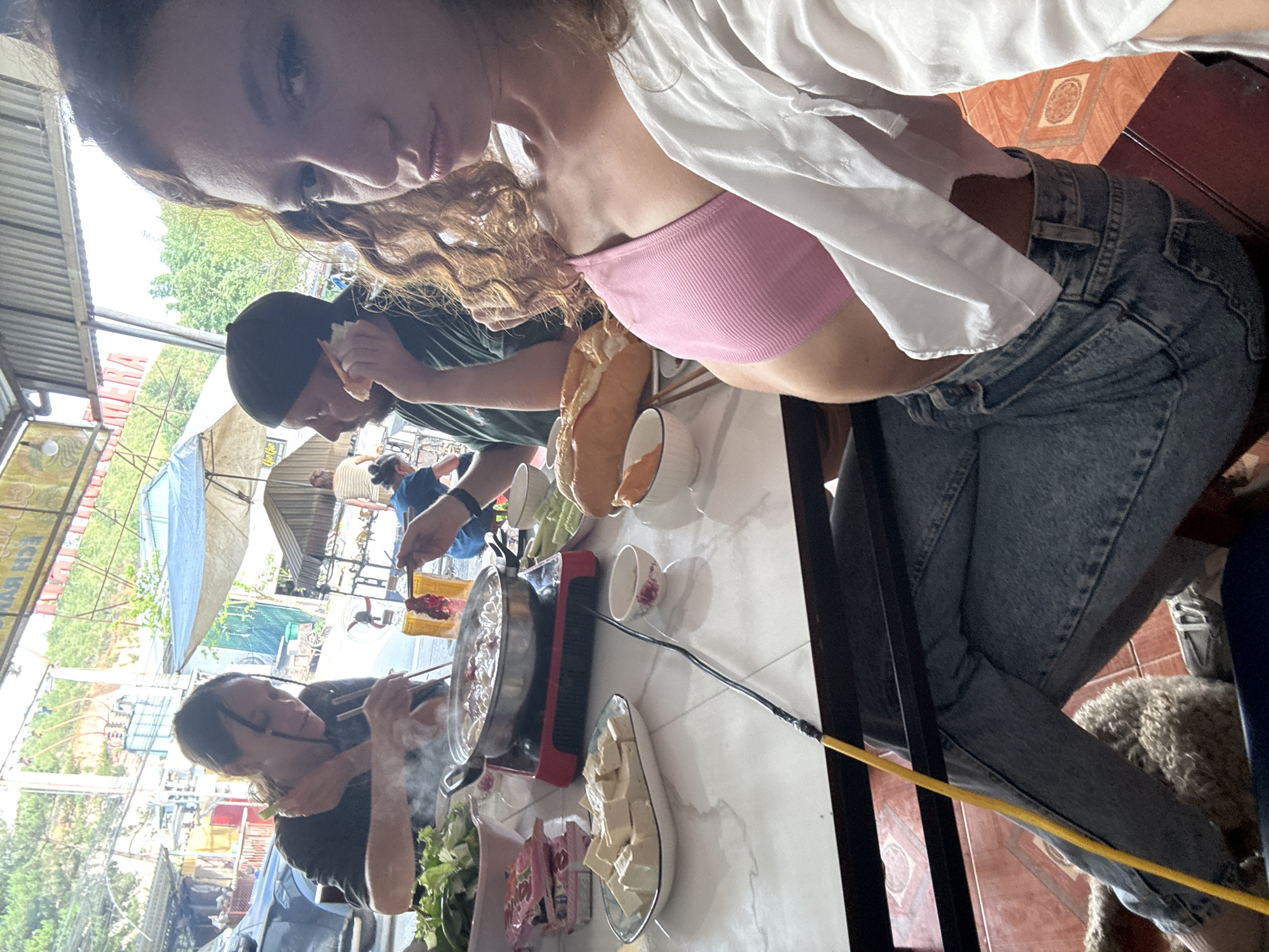 The image shows a group of people eating hot pot at a street-side restaurant in Púng Luông, Vietnam. A woman with curly hair and a pink top takes a selfie, capturing a table filled with food, including a hot pot burner, vegetables, tofu, bowls, and baguettes. Two other people are visible at the table. The restaurant is outdoors with tables covered by blue tarps and corrugated metal roofs, common for street-side establishments in Vietnam. The background shows a typical street scene, with utility poles, buildings, and other restaurant fronts.