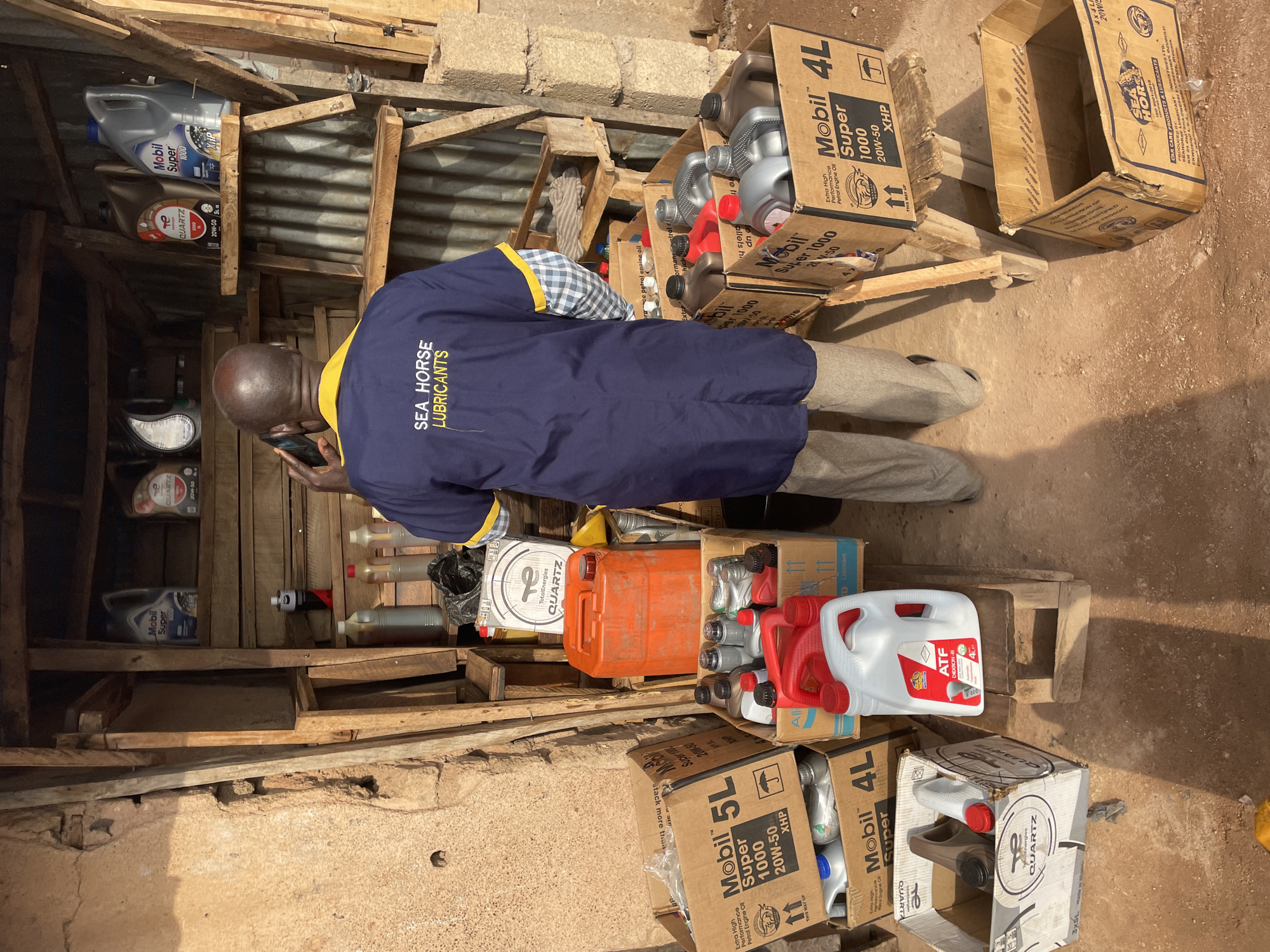 The image depicts an outdoor scene in what appears to be a small lubricant and motor oil retail location. A man, possibly the shopkeeper, is standing and looking at his mobile phone amidst a collection of motor oil products. He is wearing a navy blue work shirt with the text "SEA HORSE LUBRICANTS" printed on the back.
The products are stacked and arranged in cardboard boxes and on wooden pallets. Brands visible include Mobil and TotalEnergies Quartz. Various sizes of oil containers are present, including 4L and 5L bottles, as well as other containers of varying sizes. The products are primarily stacked on the ground. Some of the containers are in their boxes, while others are loose.
The location appears to be a simple outdoor structure, with wooden supports and metal roofing partially visible. The ground is bare and appears to be dirt or soil. Based on the text identified by OCR, it is likely located in or near Barakin Lerawi, Nigeria. The scene suggests a small, informal business operation.
