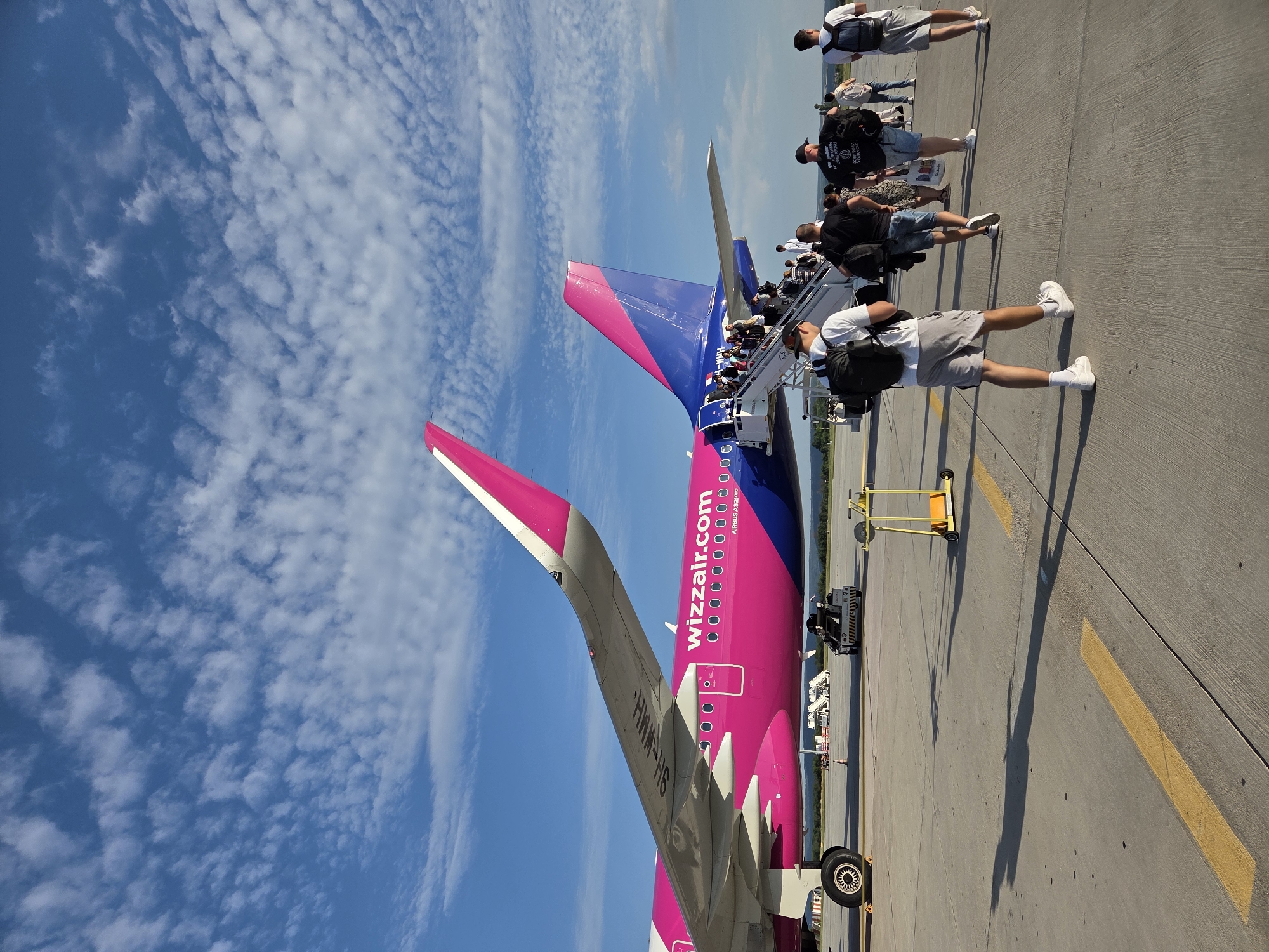 The image shows a Wizz Air airplane on a tarmac. Several people are boarding the plane using a set of stairs. The airplane is predominantly pink and blue, with "wizzair.com" printed on the fuselage. The sky is blue with scattered white clouds, suggesting it is daytime with fair weather. The ground is paved, with yellow lines indicating parking or taxiing areas. In the background, there are some trees and buildings, possibly indicating an airport environment. The people appear to be passengers, dressed in casual clothing. Some are carrying backpacks. Their body language suggests they are in the process of boarding or waiting to board the aircraft. The overall scene depicts a typical airport boarding operation on a clear day. The visible text on the aircraft is "wizzair.com" and "AIRBUS A321 NEO". The registration number of the aircraft is partially visible as "HA-M...", likely "HA-MHM". The location context provided is Aksakovo, Bulgaria.