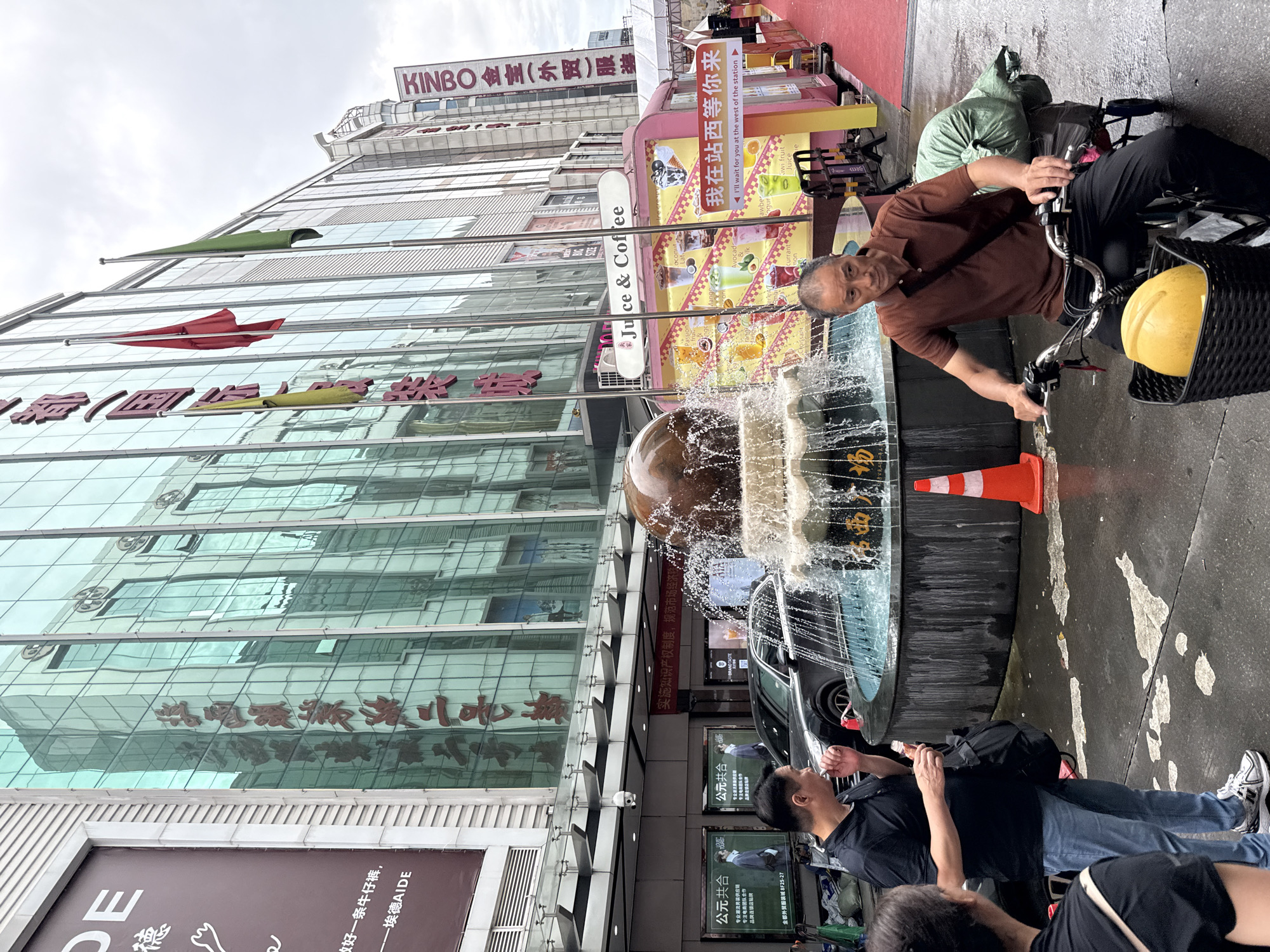 In Yuexiu District, China, a man with graying hair, wearing a brown polo shirt and black pants, rides an electric bicycle. The bicycle has a black basket on the front containing a yellow helmet and a green bag is strapped to the rear rack, obscuring the bicycle's frame. Behind him, a large, circular fountain is active, with water spraying upwards from a brown spherical object at its center. An orange traffic cone is placed to the right of the fountain. In the foreground, to the left, two individuals stand facing the fountain; one, a man in a black polo shirt and blue jeans, holds an ice cream cone, while the other, a woman in a black shirt, is partially visible. The background features a modern building with a glass facade reflecting the sky and surrounding structures. A pink food stall with colorful images of drinks and Chinese text is situated near the fountain. A red carpet extends along the right side of the scene.