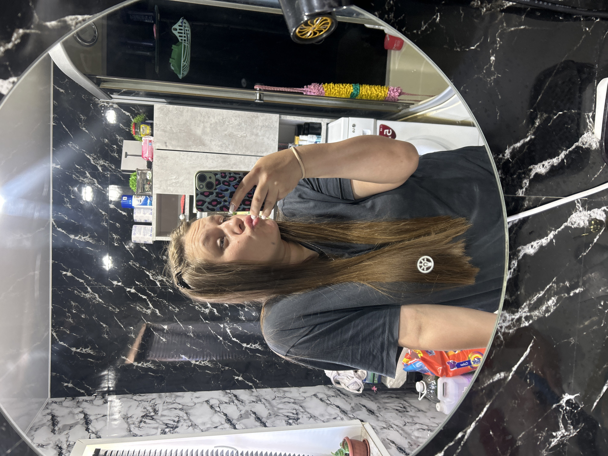 A young woman with long, brown hair is taking a mirror selfie in a bathroom. She is wearing a dark gray t-shirt and has her lips pursed. Her hair is partially braided at the front, and a small, white charm resembling a peace sign is attached to a strand of her hair. She is holding a smartphone with a leopard-print case.
The bathroom appears to have black and white marble-patterned tiles on the walls and a dark countertop. Visible in the reflection are various bathroom items, including bottles of cleaning products, a washing machine, a shower curtain, and a hairdryer. The overall impression is of a typical home bathroom. The image is undated and without any specific temporal or geographical context within Olkhovka, Russia.