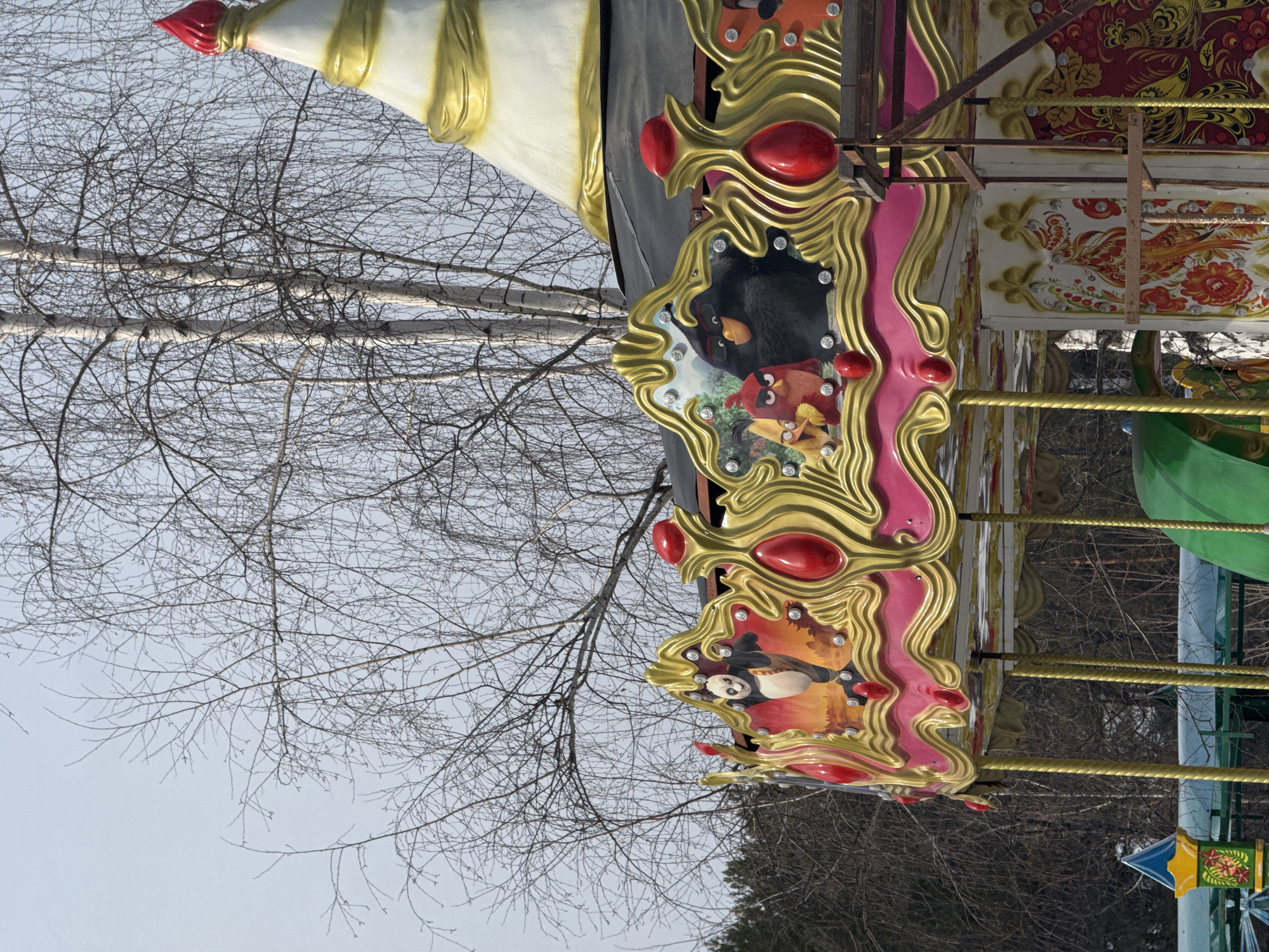 An outdoor amusement park carousel is depicted in a stationary state, likely in the city of Chaikovsky, Russia. The carousel features a conical white roof with spiraling gold banding, topped by a pointed red finial. Below the roof, a prominent decorative frieze is visible, characterized by an undulating gold frame adorned with large red ovoid jewel-like elements and smaller white circular lights. Within this frieze, panels display cartoon characters, specifically Po from "Kung Fu Panda" in an action pose on one panel, and multiple "Angry Birds" characters (Red, Chuck, and Bomb) on another. The main body of the carousel is decorated with white panels featuring intricate red and orange folk-art patterns of birds and floral motifs. Twisted gold-colored vertical support poles descend from the frieze. Wooden scaffolding-like structures are present on the right side of the carousel, suggesting maintenance or ongoing construction/deconstruction. The background consists primarily of bare deciduous trees, including multiple white-barked birch trees, set against a light grey, overcast sky. Evergreen trees are visible in the deeper background to the left. Partial elements of other amusement park rides, including a blue and yellow kiosk-like structure and green ride components, are visible in the lower foreground. No individuals are present in the scene.