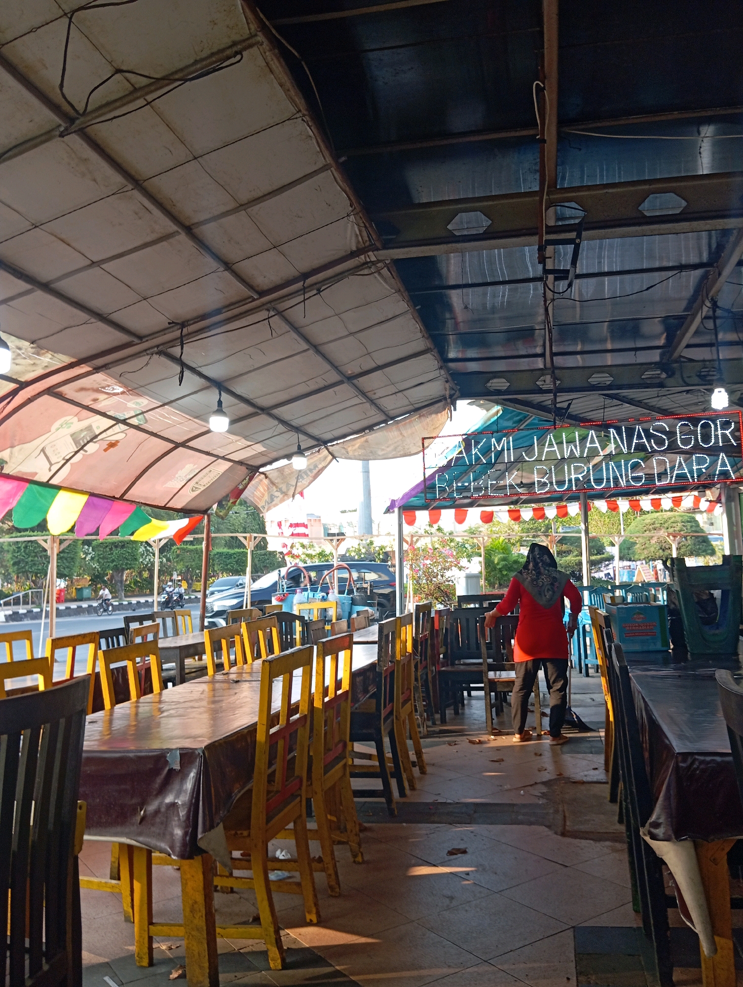 The image captures an outdoor dining area of a restaurant in Semarang, Indonesia, likely during the daytime as indicated by the sunlight casting shadows. The scene features rows of wooden tables covered with dark plastic tablecloths, surrounded by brightly colored chairs – primarily yellow with some red accents. A woman wearing a red top and a headscarf is seen standing in the middle of the seating area, appearing to sweep the floor. Above the dining area, a translucent, multicolored canopy covers the space, with lighting fixtures attached. A neon sign with red and blue lettering is visible, displaying text that appears to be in Indonesian, likely related to menu items such as "NAS GOR" (fried rice) and "BEBEK BURUNG DARA" (dove/pigeon). In the background, traffic can be seen on a road, with trees and some buildings forming the city landscape. The ground is tiled, and fallen leaves are scattered across it, suggesting a somewhat untidy or natural outdoor environment.