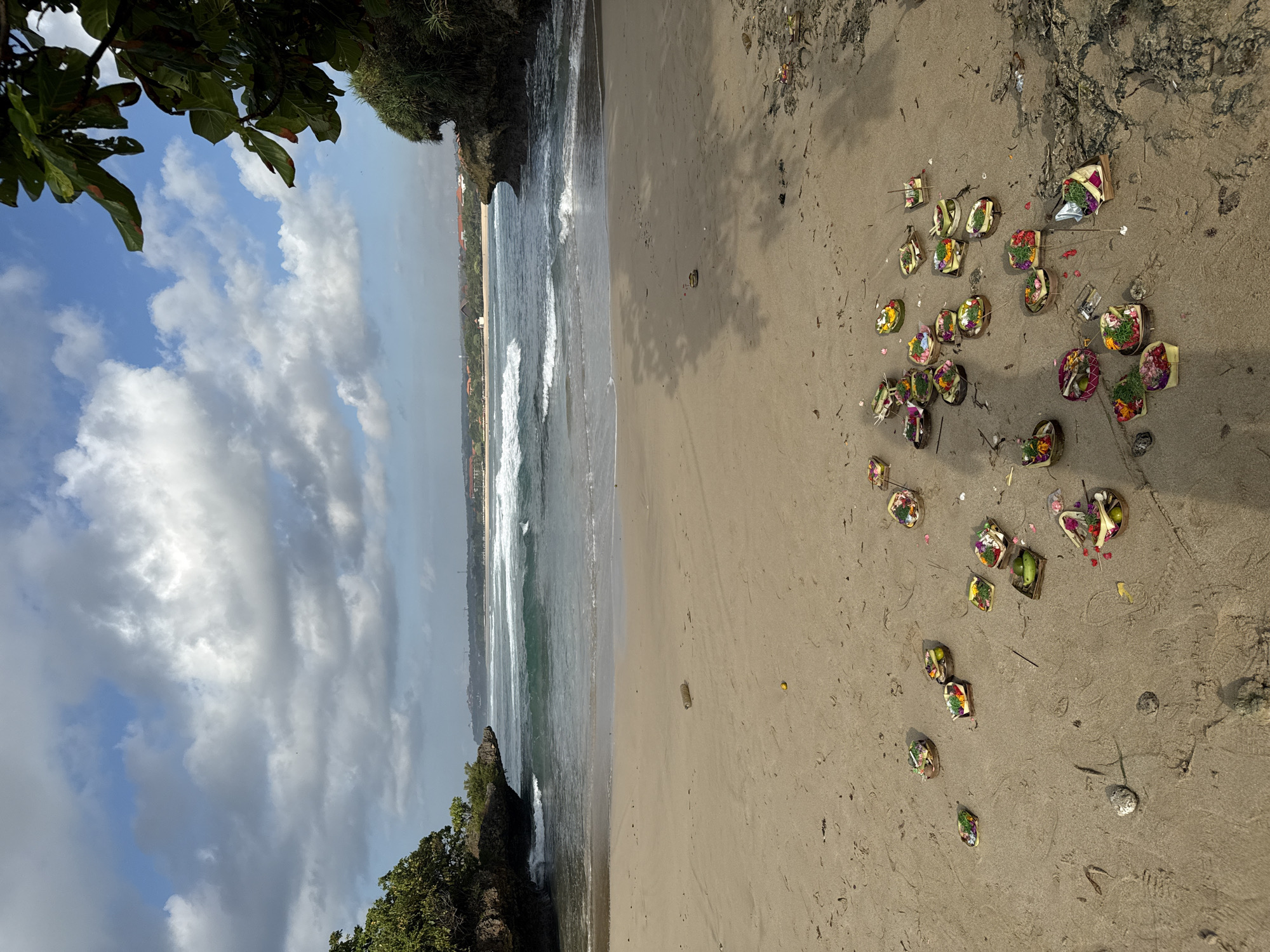 This is a medium shot, eye-level, vertical photograph taken on a beach in Kuta Selatan, Indonesia. The foreground is dominated by a sandy beach with numerous offerings scattered across it. These offerings are small woven baskets filled with colorful flowers, fruits, and incense sticks. Some of the baskets appear to be partially decomposed, with organic matter visible. Among the offerings, there are a few rocks and some debris, including a plastic bottle and a discarded piece of shoe on the far right. The sand has tire tracks and footprints.
In the middle ground, gentle waves are breaking on the shore, creating white foam. The water is a clear turquoise color. On either side of the beach, rocky outcrops covered in green vegetation frame the view of the ocean.
The background shows a distant coastline with buildings and more vegetation. The sky is a bright blue with scattered white and grey clouds, indicating a partly cloudy day. The overall lighting suggests it is daytime, with shadows cast on the sand from the surrounding foliage.