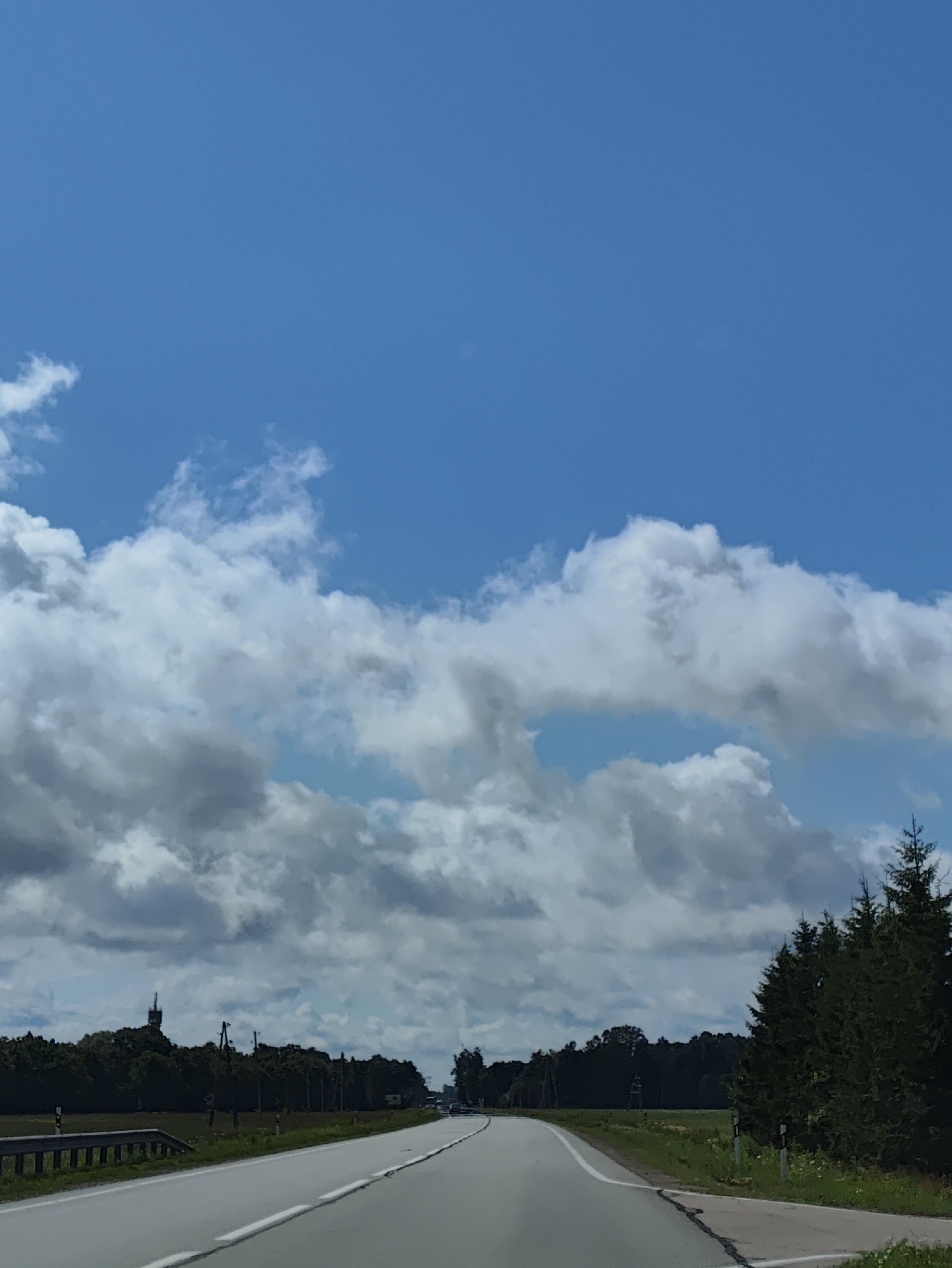 The image depicts a view of a highway leading through a rural landscape in Svētciems, Latvia. The sky is a mix of bright blue and dramatic, billowy clouds, suggesting dynamic weather conditions. The road itself is paved with white lane markings and extends into the distance, curving slightly to the right. On the left side of the road, a metal guardrail runs alongside a grassy shoulder. To the right of the road, a field of green grass and wildflowers is visible, bordered by a dense line of dark green evergreen trees. In the far distance, silhouetted against the sky, a tall, slender structure, possibly a radio tower or a church spire, stands amidst the trees. The overall impression is one of a serene, yet vast, natural environment traversed by a modern roadway.