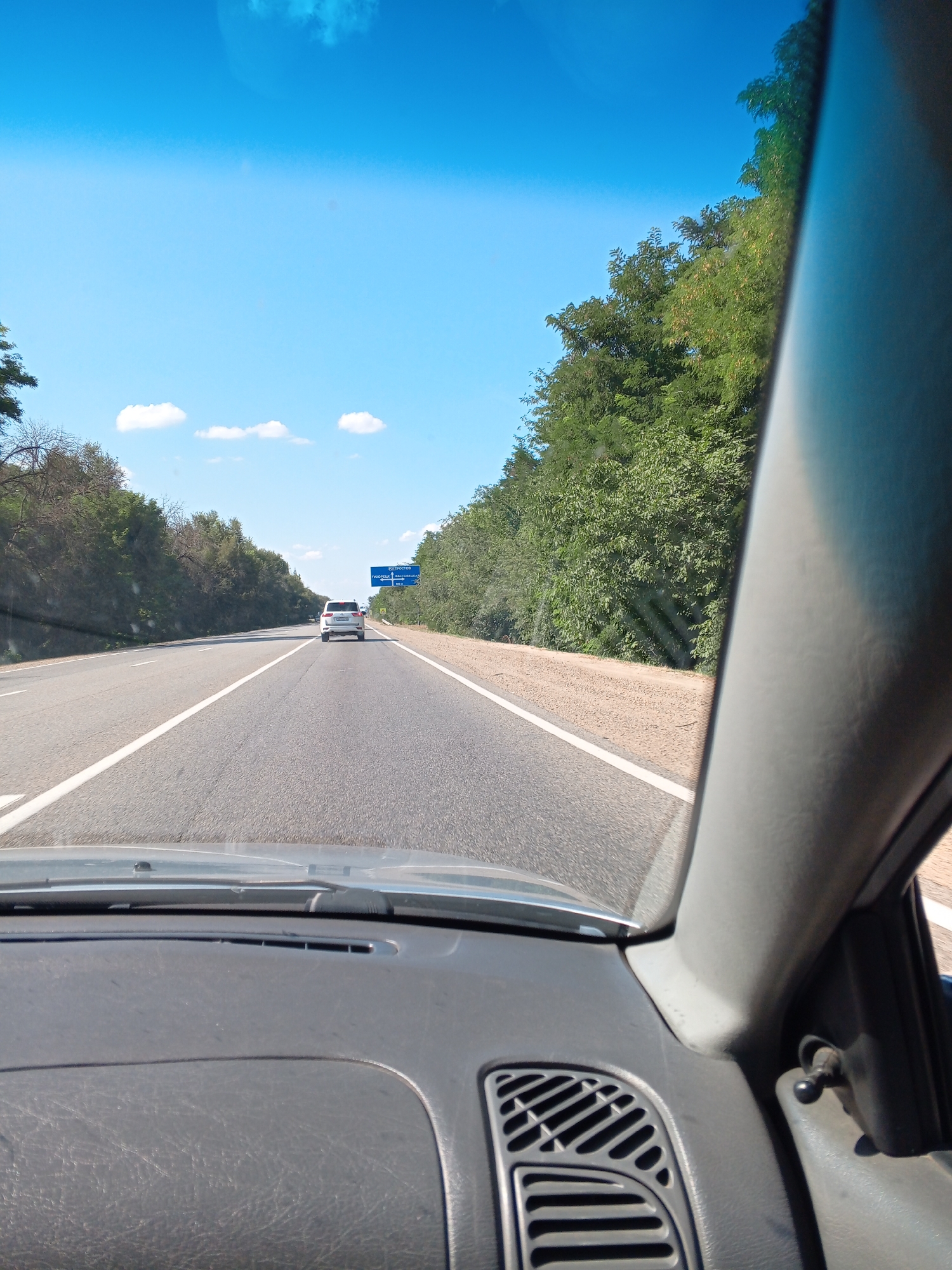 This is a frontal view from inside a vehicle traveling on a highway. The road ahead has multiple lanes and is lined with trees on both sides. The sky is clear blue with a few scattered clouds. In the distance, a white SUV is visible, and a blue road sign is also present. The interior of the vehicle, including the dashboard and a side pillar, is visible in the foreground. The image depicts a sunny day on a highway.

The road sign in the image indicates directions to Tikhoretsk, Russia. Specifically, the sign displays "ТИХОРЕЦК" (Tikhoretsk) and "Г. ТИХОРЕЦК, ул. Дзержинского" (City of Tikhoretsk, Dzerzhinsky Street). This suggests the vehicle is on a highway leading to or from the city of Tikhoretsk.