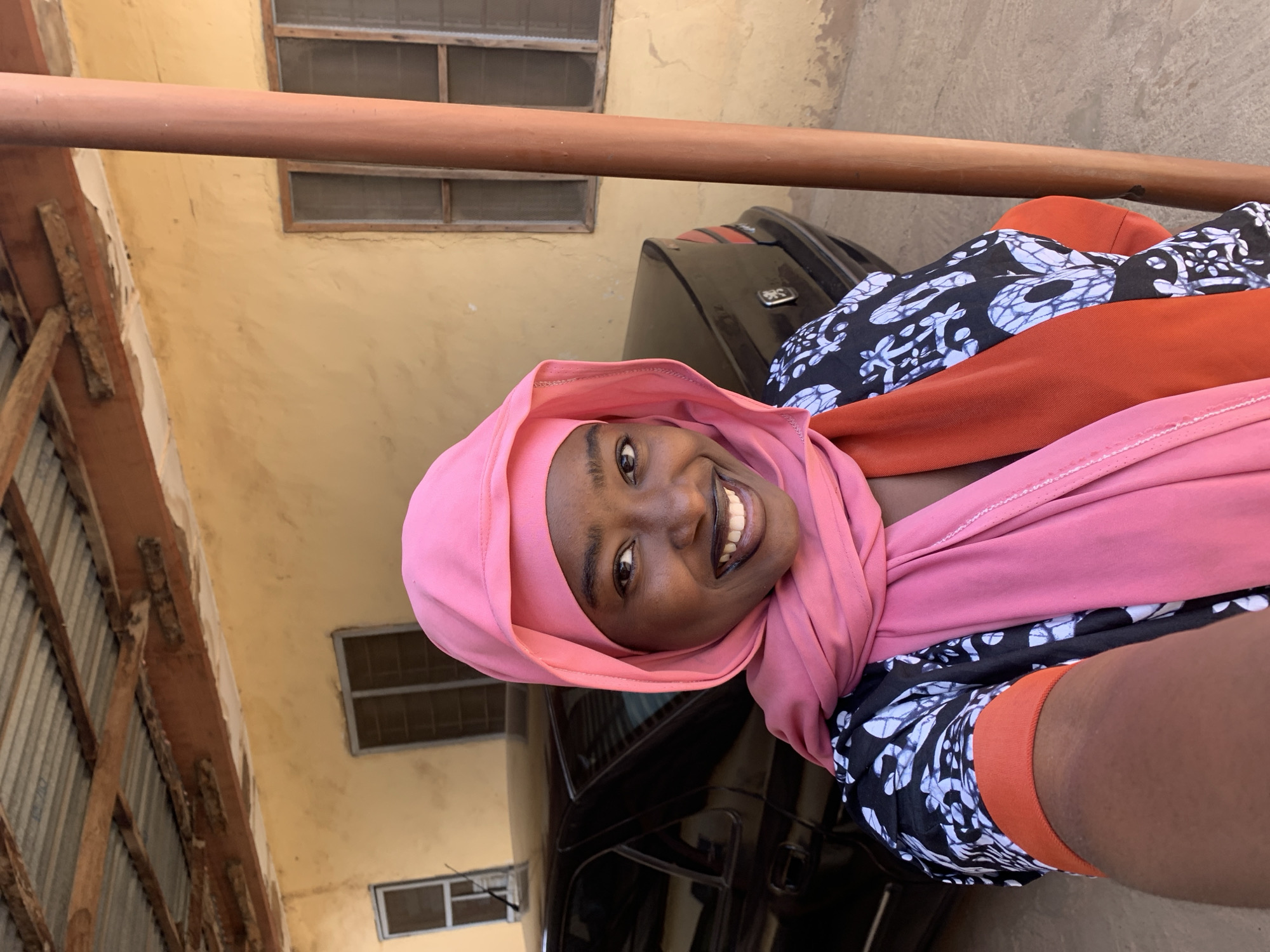 A woman with dark skin is smiling at the camera while wearing a pink hijab and a patterned black and white shirt with orange trim. She is positioned near a black car with a reflective surface. Behind her, a beige building with rectangular windows is visible, and a wooden structure is on the left. The location is indicated as Rafin Par, Nigeria, based on the prompt instructions.