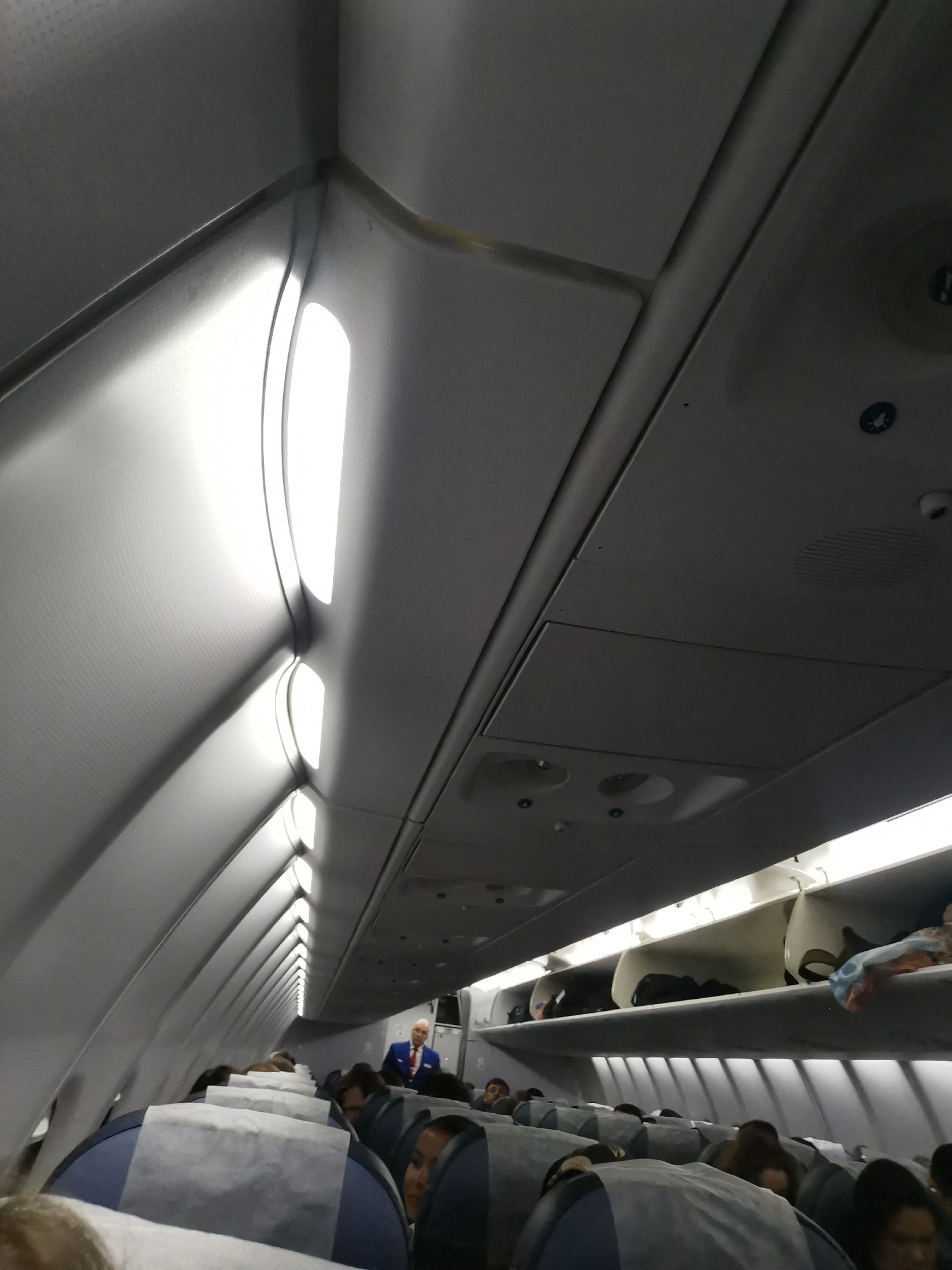 The image shows the interior of an airplane cabin. Passengers are seated in rows of seats with blue and gray upholstery. Overhead luggage compartments are open and contain bags and belongings. A flight attendant wearing a blue suit and red tie is visible at the front of the cabin. The cabin ceiling features lights and ventilation systems. The location in Новое Мартьяново, Russia is not discernible from this image.