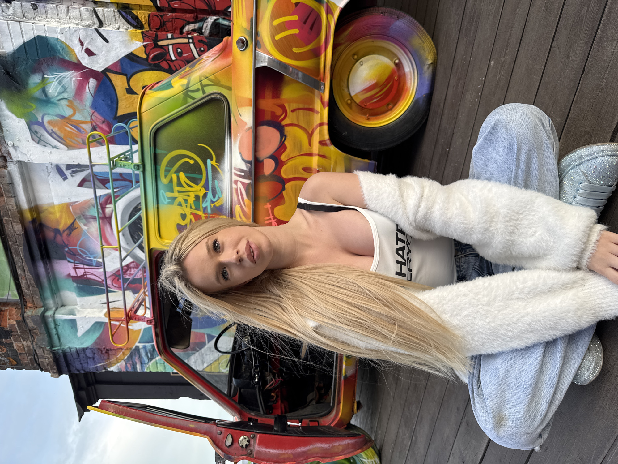 A young woman with long blonde hair sits next to a brightly painted vintage car. The car is covered in graffiti, with vibrant colors like yellow, orange, red, and blue. The woman wears a white crop top with black trim, a fluffy white shrug, and light-wash jeans. Her shoes are sparkly silver sneakers. She is looking towards the camera with a neutral expression. The car appears to be a small, older model, possibly a Trabant, with its door open. The background is a wooden deck and a brick wall also covered in graffiti. The lighting suggests it is daytime, and the weather is likely clear. The scene appears to be an urban setting, possibly an outdoor exhibition or a unique street art installation. The text on the woman's shirt reads "HATE EVERYONE". The location is identified as Saint Petersburg, Russia.