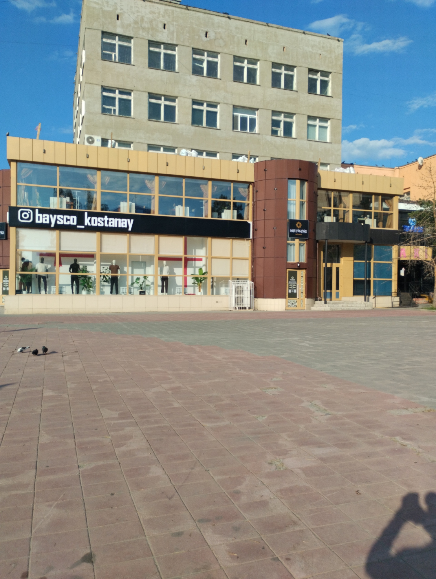 A concrete building stands tall against a bright blue sky with scattered clouds. Below it, a modern storefront with large glass windows displays mannequins dressed in clothing. The windows are framed with a light-colored material. A sign above the windows reads "@baysco_kostanay" in white lettering on a black background. To the right of this storefront, another section of the building features a rounded brown facade with more glass windows and an entrance. A sign with Cyrillic text and a stylized sunburst logo is visible above this entrance. The ground in front of the building is paved with red and gray square tiles. Several pigeons are visible on the tiled ground, with one white and gray pigeon standing and two darker pigeons near it. In the lower right corner, the shadow of a person holding a camera is cast on the pavement. The image appears to be taken in the city of Kostanay, Kazakhstan, based on the visible signage.