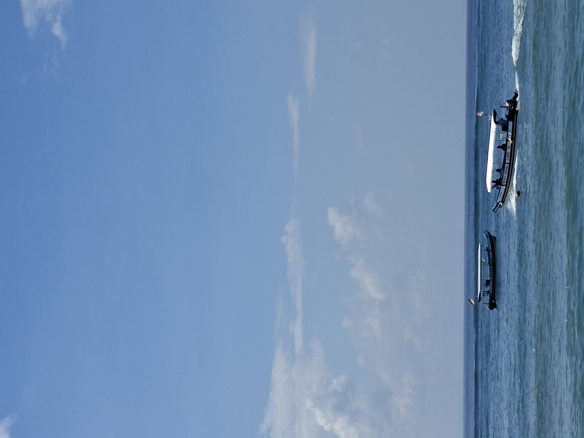 The image captures a vibrant daytime scene of the ocean under a bright blue sky, with two boats traversing the water. The horizon line is visible, separating the expansive sky from the sea.
The sky is predominantly clear and a uniform light blue, with a few scattered, wispy white clouds adding subtle texture. The ocean is a deep blue-green, appearing slightly choppy with small waves.
In the mid-ground, towards the right side of the frame, two elongated boats are visible on the water. The boat closer to the viewer is larger, with a white hull and a dark canopy covering its seating area. Several individuals are seated within this boat, suggesting it is carrying passengers. It appears to be moving, as indicated by a distinct white wake trailing behind it. On the side of this boat, text is visible, which reads "PERAHU WISATA" (Indonesian for "TOURIST BOAT"), followed by what appears to be "PENGAYU" and some numbers.
Further back and slightly to the left of the larger boat is a second, smaller boat. This boat has a dark hull with a white stripe along its side. Fewer people are discernible on this vessel, possibly one or two. It seems to be either stationary or moving at a slower pace compared to the front boat, with less apparent wake.
The overall impression is one of a sunny day, likely late morning or early afternoon, suitable for marine activities. The presence of "TOURIST BOAT" text and multiple passengers suggests a tourism-related activity. Given the context of Kuta Selatan, Indonesia, this scene is consistent with coastal activities and transportation common in that region.