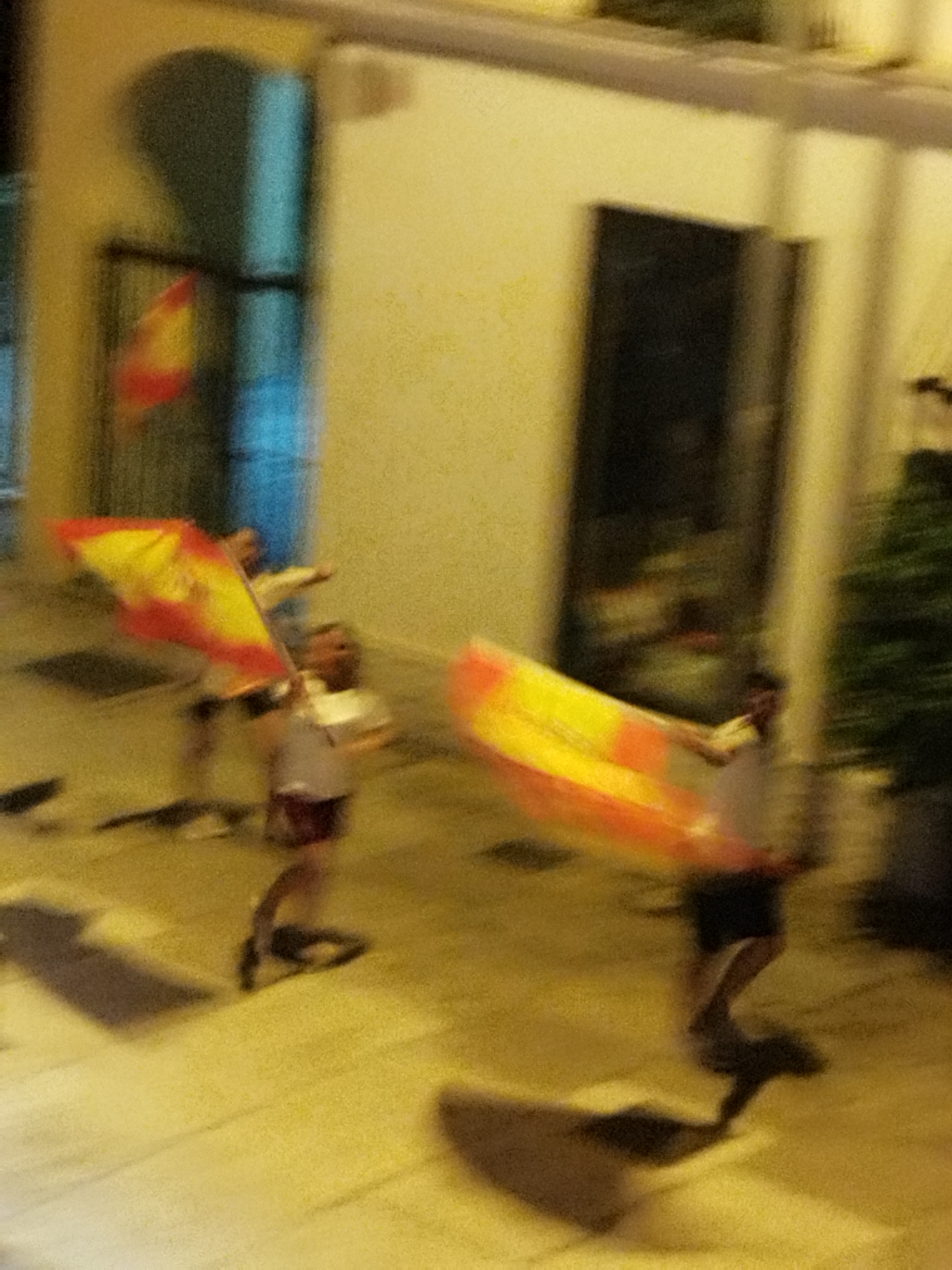 The image shows a blurry outdoor scene at night, with several people holding and waving Spanish flags. The setting appears to be a city street or plaza, possibly during a celebration or event. The lighting is dim and creates long shadows. In the background, there is a building with yellow walls and a dark archway. The motion blur suggests movement and activity. While the specific location in Jerez, Spain, cannot be definitively identified, the presence of Spanish flags suggests a Spanish context. The overall impression is one of festivity and national pride.