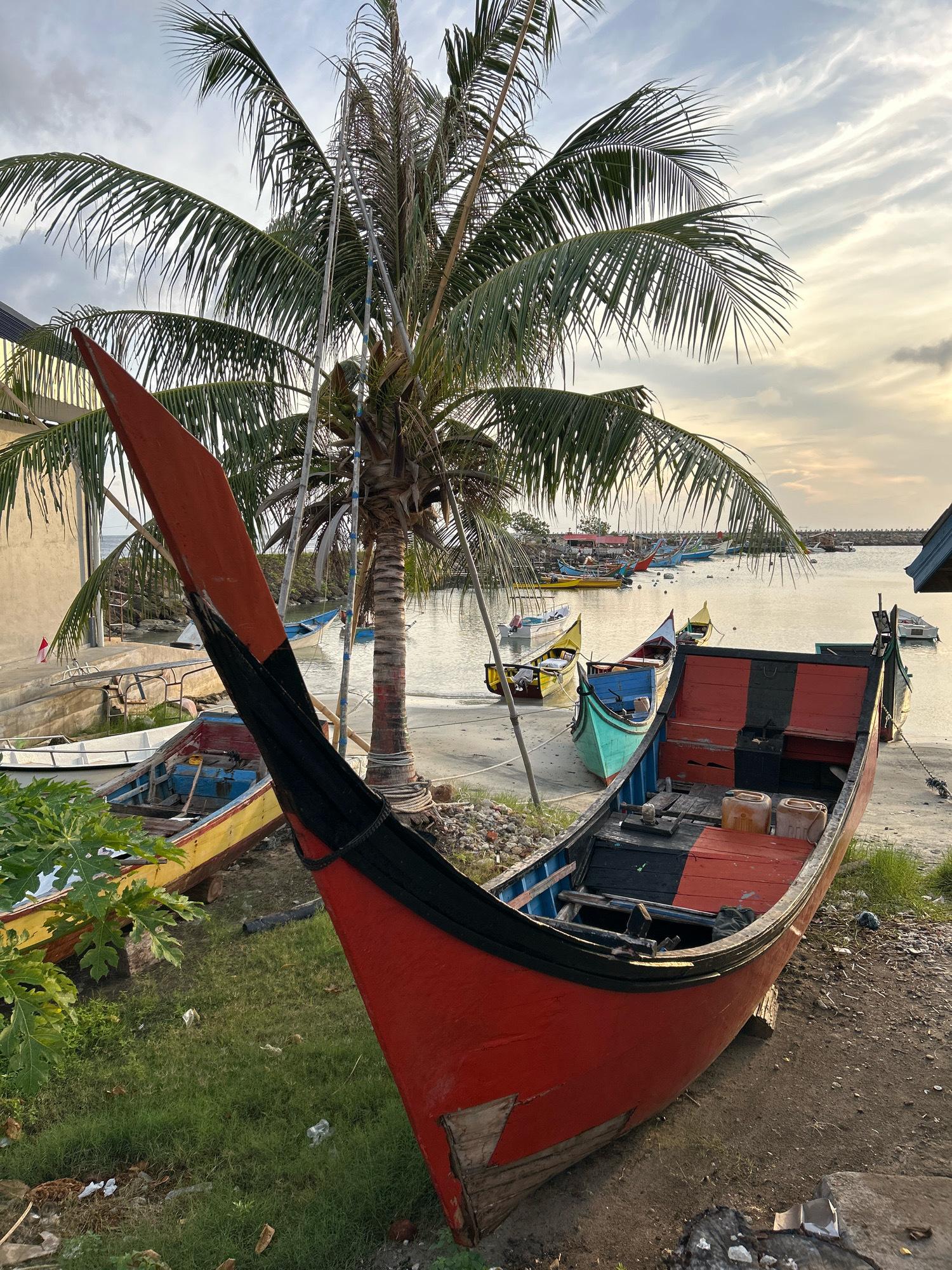 A coastal scene in Sabang, Indonesia, features a harbor with numerous fishing boats docked or beached. In the foreground, a large, weathered red and black fishing boat is prominently positioned on the sandy shore. It shows signs of age and use, with peeling paint and visible wooden planks. Two orange jerrycans are stored inside the boat. Behind this main vessel, a cluster of smaller boats in various colors, including yellow, blue, and green, are also beached or moored in shallow water. A palm tree with lush green fronds stands to the left of the red boat, its trunk wrapped with some ropes and metal poles. The background reveals more boats in the distance, a breakwater, and a hazy sky with soft clouds, suggesting either early morning or late afternoon light. A building with a pale facade is visible to the left, partially obscured by the palm tree. The overall environment is a working harbor with the characteristic elements of a tropical coastal community.