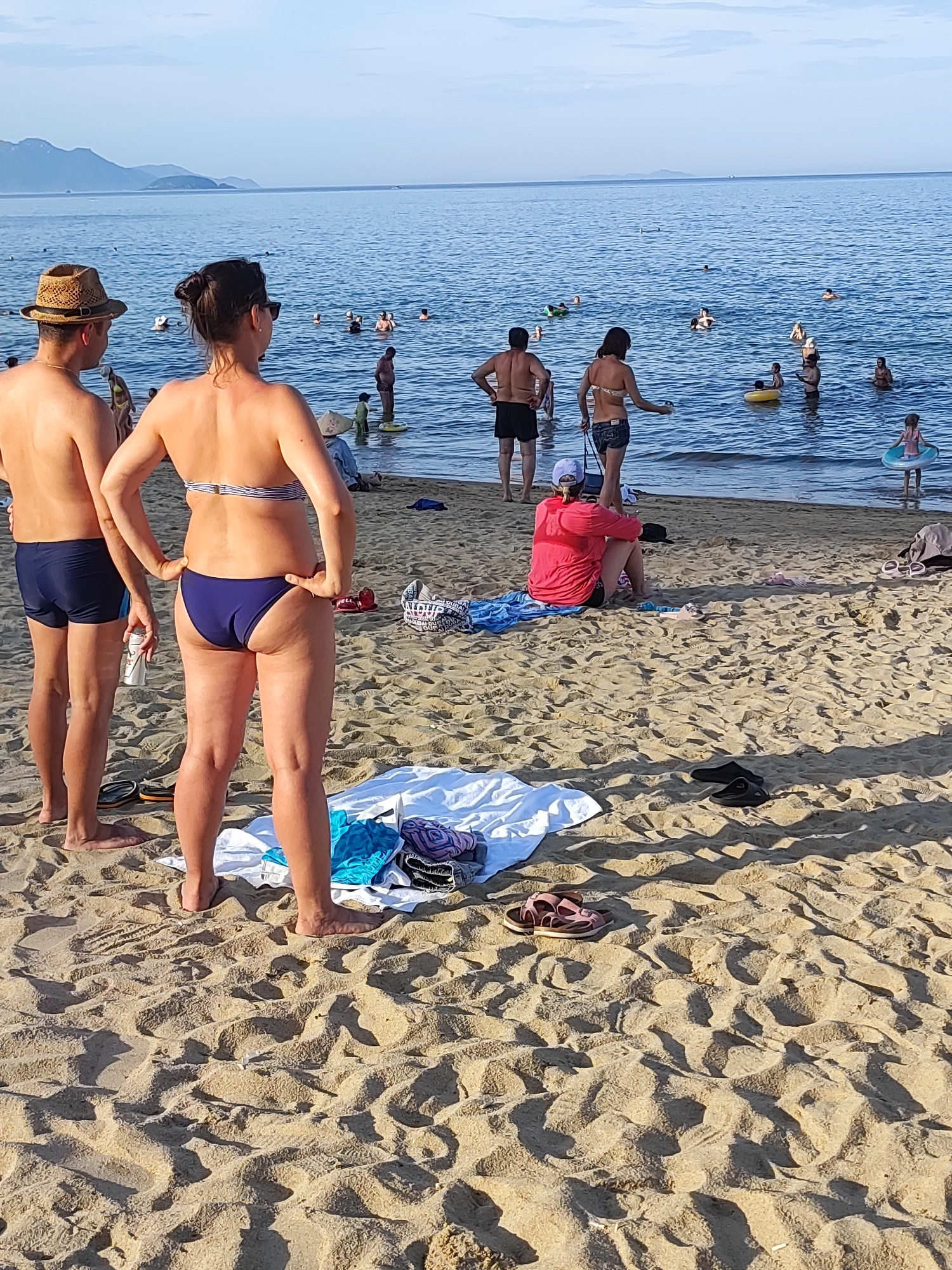 The image depicts a crowded beach scene on a sunny day, likely in Nha Trang, Vietnam, given the presence of a conical hat worn by one of the individuals and the general tropical atmosphere. In the foreground, two individuals are standing on the sand, facing away from the camera, with the ocean and other beachgoers in the background. The man on the left is wearing a straw hat and dark blue swim trunks, while the woman next to him is in a bikini and shorts. The sand is covered with footprints, some towels, and sandals.
The ocean is filled with people swimming, wading, and enjoying various water activities, with several inflatable rings visible. The water is a clear blue, and the sky above is a pale blue with some distant mountains on the horizon. The overall impression is a lively and typical beach day.