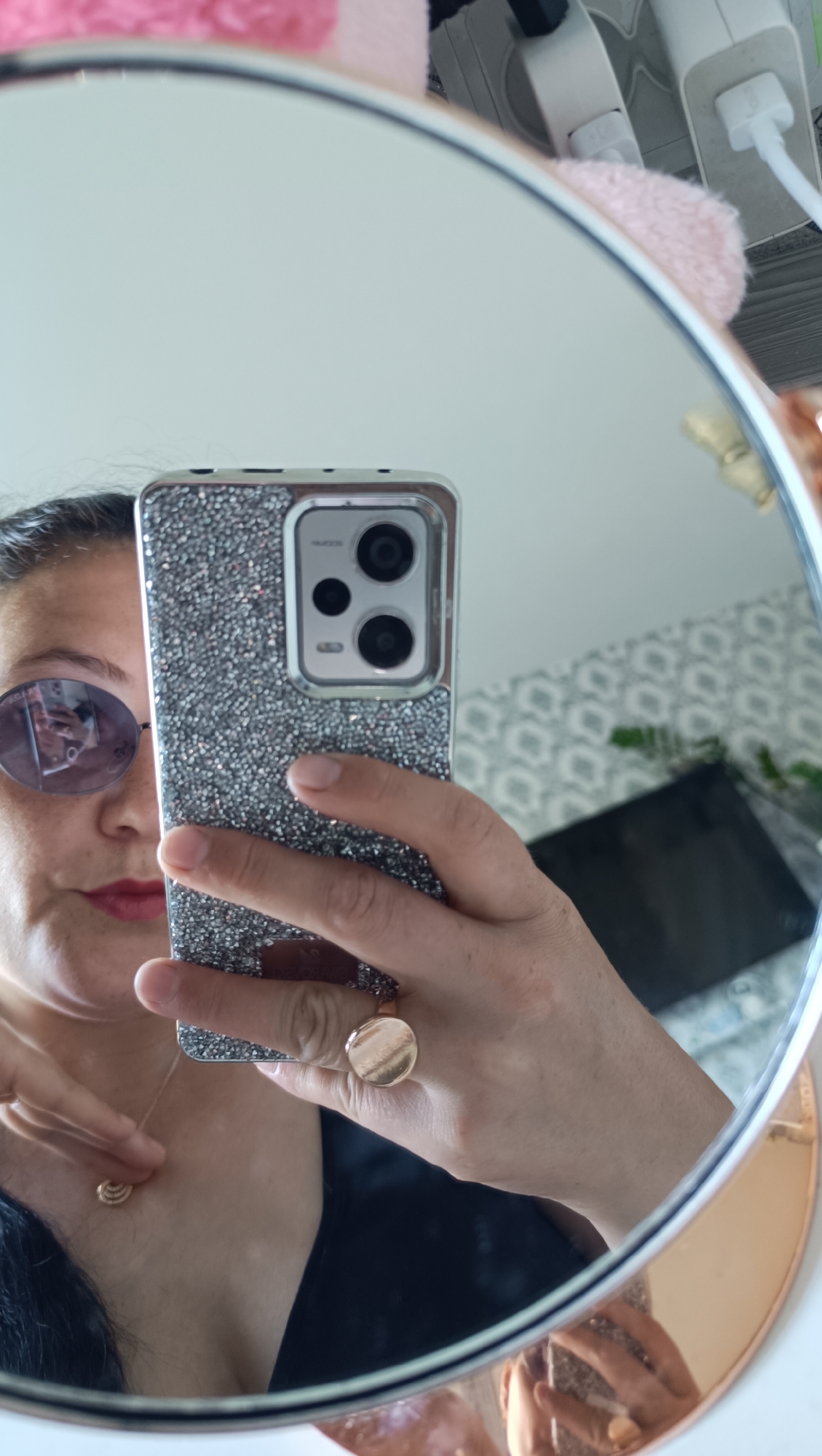 In a mirrored reflection, a woman is taking a selfie. She is wearing sunglasses and has red lipstick. On her left hand, she wears a large, circular gold ring. On her neck, she wears a delicate gold necklace with a shell-shaped pendant. She is holding a smartphone with a silver, rhinestone-encrusted case. The background of the reflection shows a wall with a patterned wallpaper and electrical outlets. The mirror is round with a rose gold frame. The scene does not provide any information that could place it in Chaikovsky, Russia.