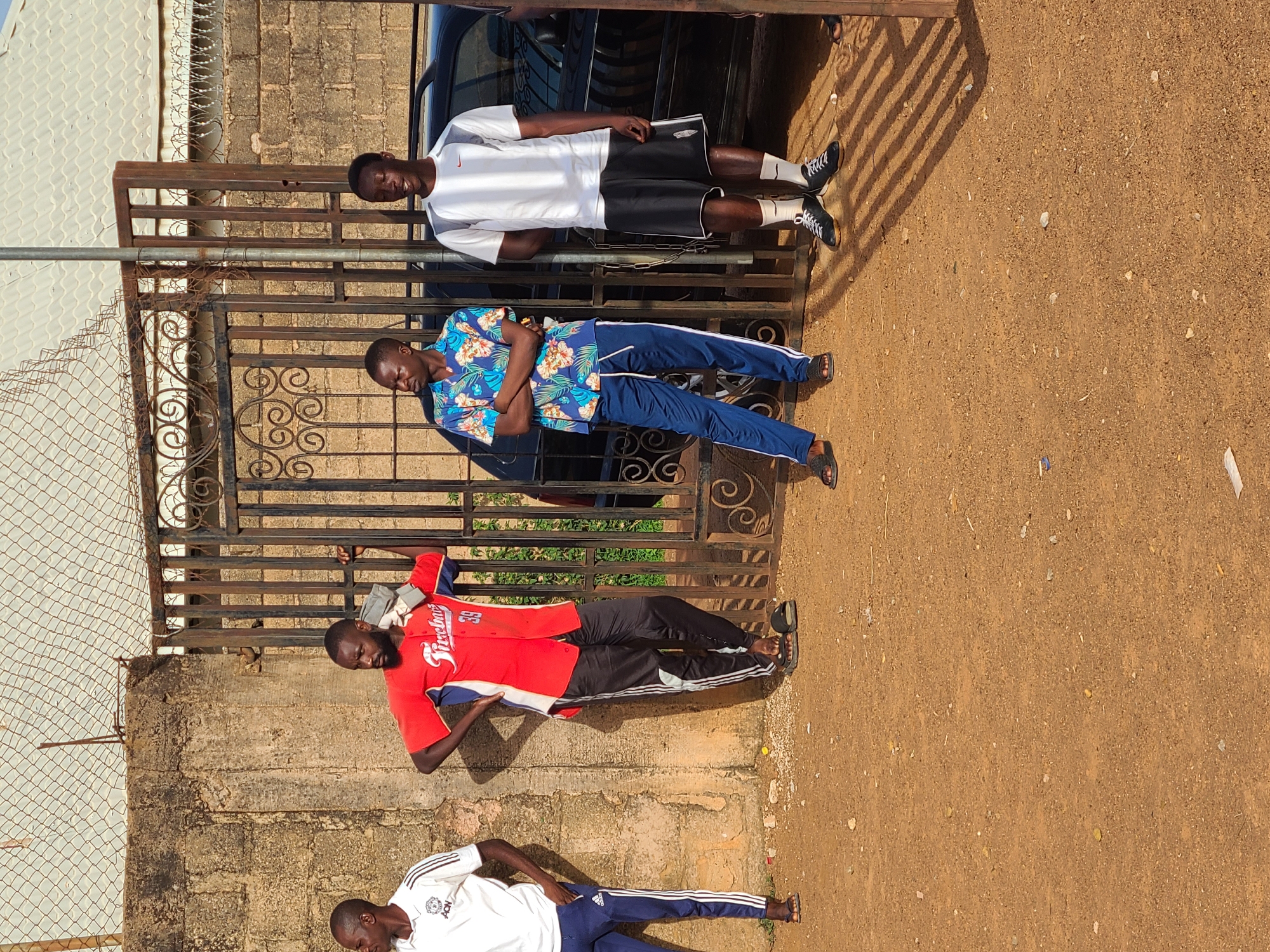 The image shows four young men in front of a gate in Jos, Nigeria. The first man is wearing a white shirt and black shorts. The second man is wearing a blue floral shirt and blue sweatpants. The third man is wearing a red shirt and black sweatpants. The fourth man is wearing a white shirt and blue sweatpants. The gate is made of metal and is located in front of a wall. The ground is dirt. A black car is parked behind the gate.