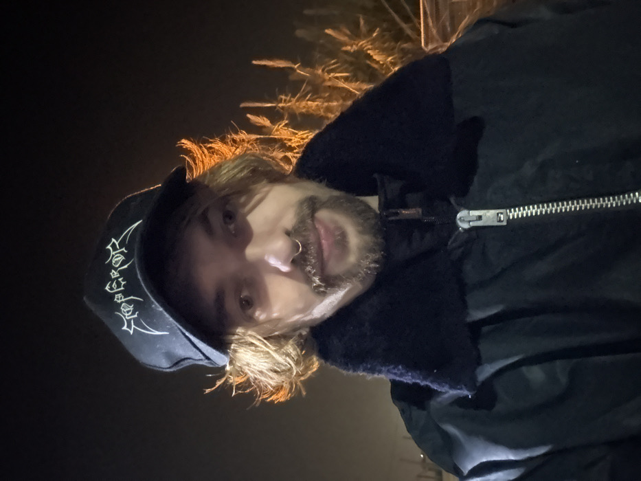 A person with blonde hair, a beard, and a nose ring poses in a low-light setting. They wear a dark baseball cap with stylized white graphics and a dark jacket with a fur collar and a visible zipper. Tall, dry grass illuminated by an unseen light source is visible behind them. The photo appears to have been taken at night or in a very dimly lit area.