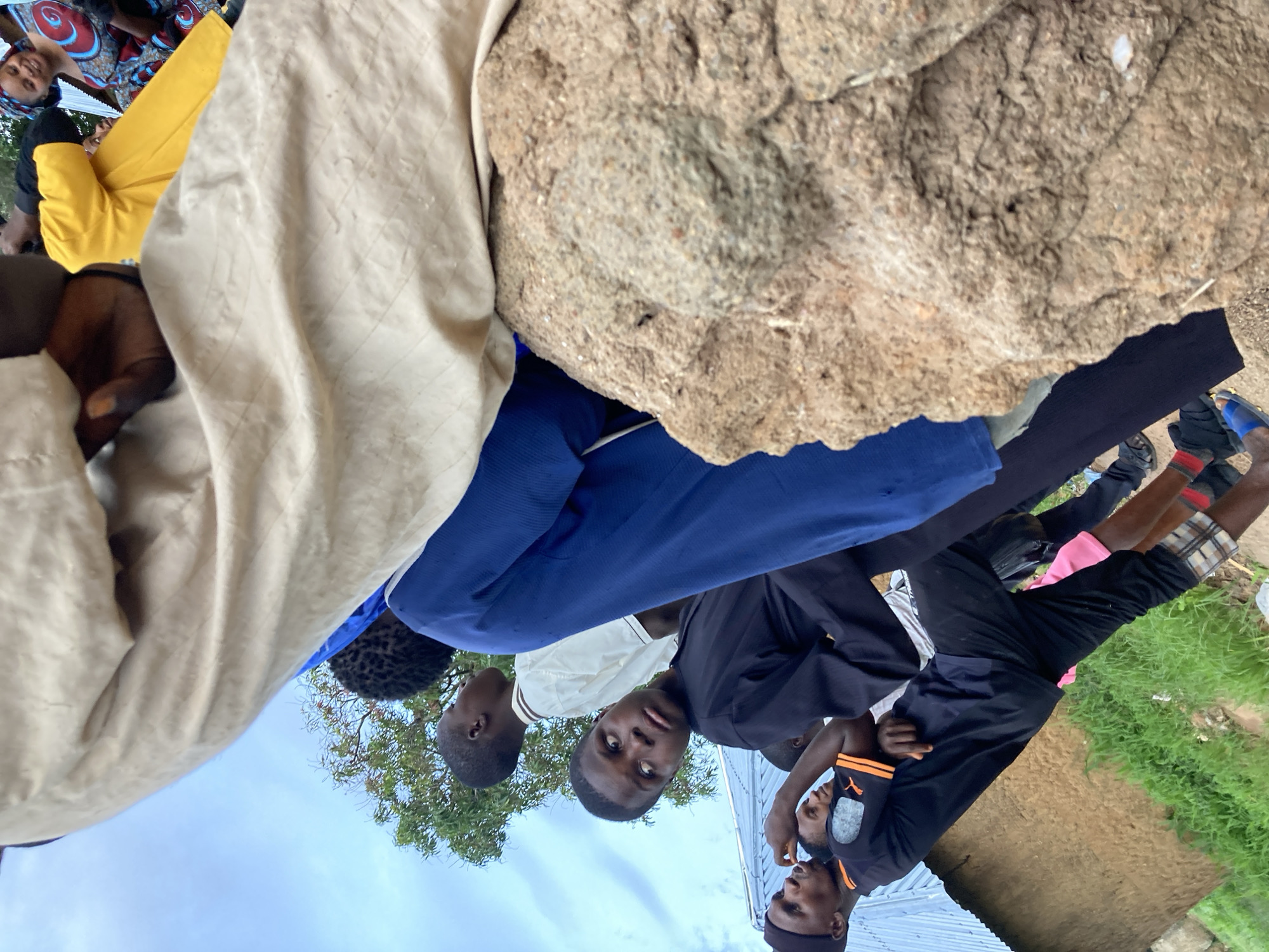 The image depicts a group of people, predominantly young males, gathered outdoors in what appears to be a residential area in Jos, Nigeria. The setting includes a simple structure with a corrugated metal roof and a mud-brick wall, surrounded by green vegetation. A large, rough, earthy mound is prominently featured in the foreground, obscuring a portion of the scene.
Several individuals are visible, with varying degrees of clarity. One young male in the foreground, wearing a black outfit, is looking towards the left with a concerned or curious expression. Another young male in a white shirt is looking upwards. Further back, a woman in a colorful patterned dress is smiling. The people are dressed in casual clothing, with some wearing traditional attire. The overall atmosphere seems informal, possibly a community gathering or a moment of pause. The sky above is partly cloudy, suggesting daylight.