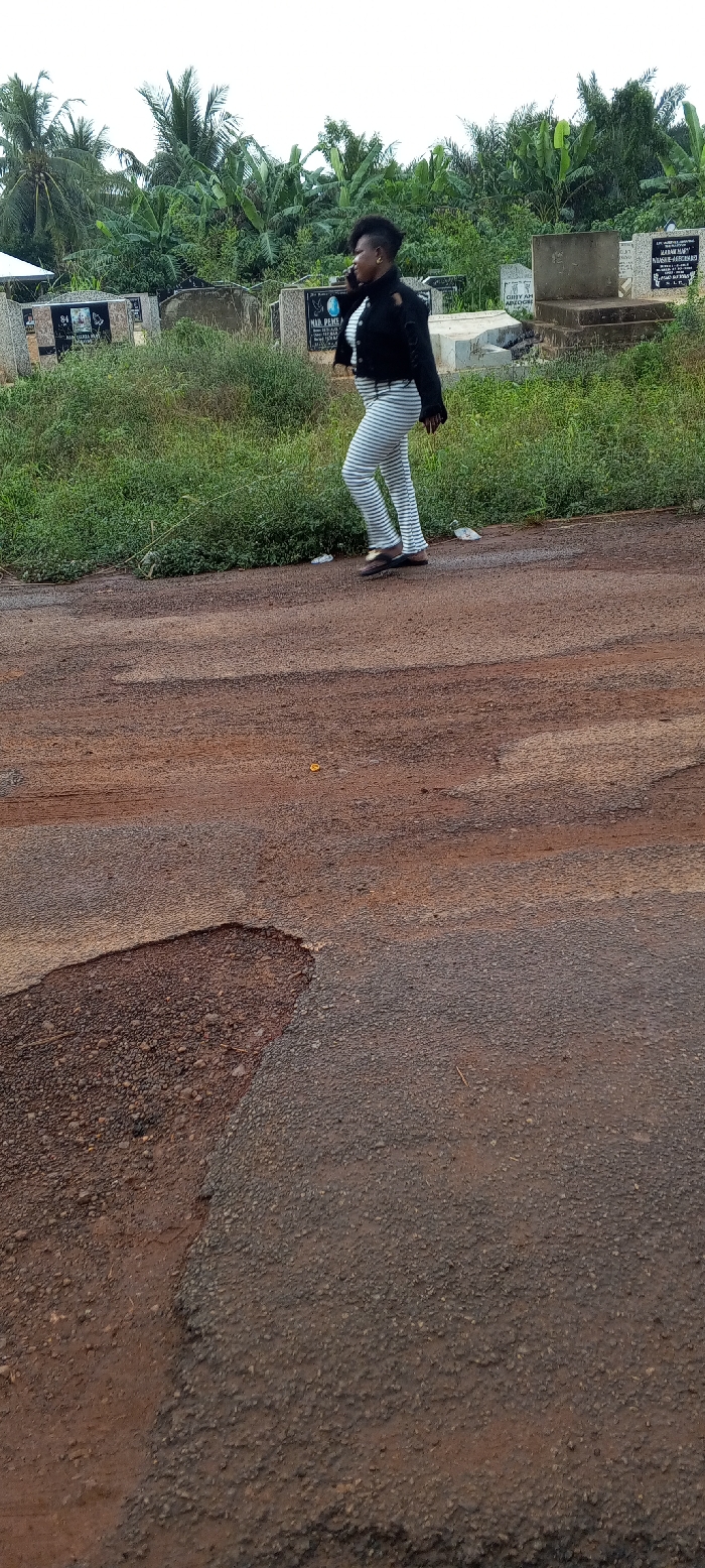 An individual walks on a partially paved road adjacent to a cemetery in Dzodze, Ghana. The person, with dark skin and a short, spiky haircut, is seen in profile moving from left to right across the mid-ground. They are wearing a black long-sleeved jacket with shoulder cutouts, horizontally striped white and grey pants, and dark flip-flops, while holding a dark mobile phone to their left ear. The road surface is degraded, featuring areas of asphalt mixed with exposed red-brown earth and potholes. To the left of the road, a strip of green grass separates it from a cemetery containing multiple concrete grave markers and tombs, some with visible inscriptions like "MR. PEACE." Dense tropical vegetation, including palm and banana trees, forms the background behind the cemetery. The sky is bright and overcast. A small yellow object and white debris are visible on the road surface in the foreground.