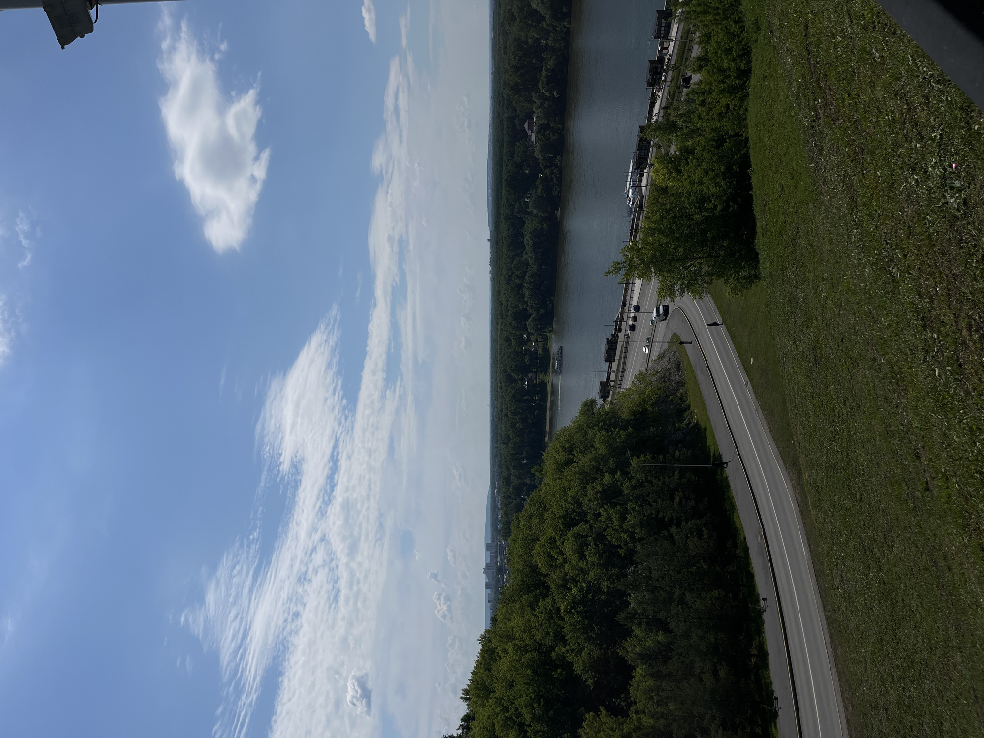 The image depicts a panoramic view from a grassy hill overlooking a wide river, likely the Belaya River, in Ufa, Russia. In the foreground, a winding asphalt road with white lane markings curves along the base of the hill, with some grass growing on the embankment. A few vehicles, including cars and scooters, are visible on the road. A single person is walking on the sidewalk adjacent to the road.
Across the river, a dense, green forest stretches as far as the eye can see, dotted with some scattered buildings. In the distance, the silhouette of a city skyline with several tall buildings is visible against the horizon. A large boat is navigating the river, and several smaller boats are docked along the riverbank. The sky is a bright blue, with scattered white clouds. The overall impression is a serene, natural landscape blended with urban elements.