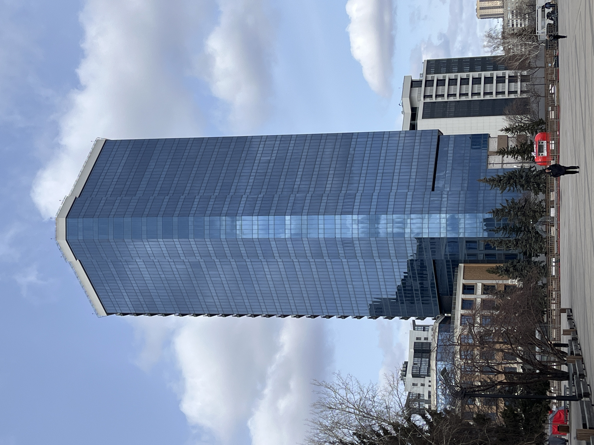 The image shows a tall, modern, glass-fronted skyscraper in Krasnoyarsk, Russia. The building is rectangular and predominantly blue-gray in color due to the reflective glass panels. The sky above has a mix of blue and white clouds. In the background and to the side of the skyscraper are other buildings, some with distinct white and dark-colored facades. Trees are sparsely scattered in the middle ground. A paved area, with benches and some individuals walking, stretches out in front of the buildings. A red snack or drink kiosk is also visible.