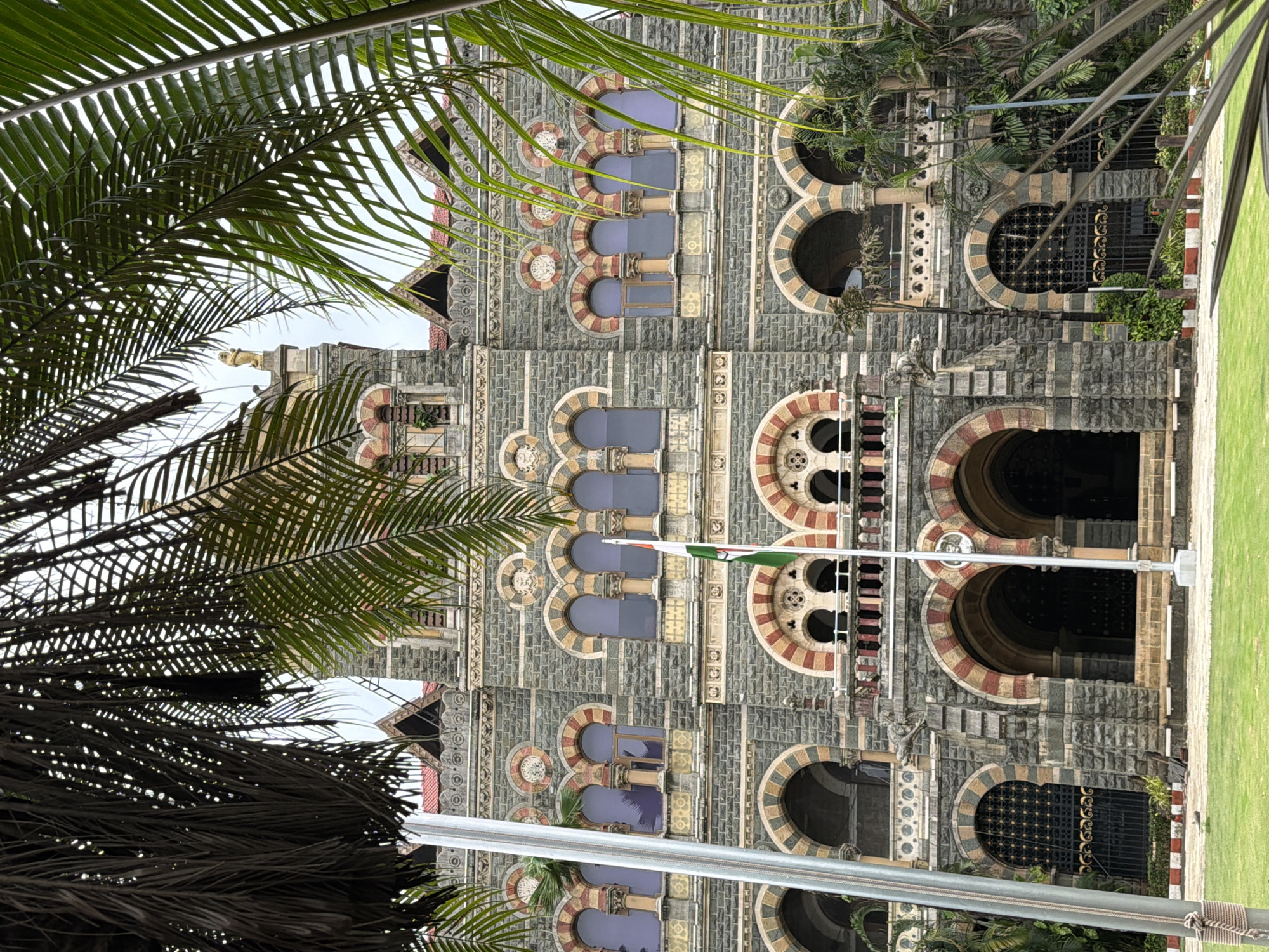 The image shows the Chhatrapati Shivaji Maharaj Terminus (CSMT), formerly Victoria Terminus, a historic railway station in Mumbai, India. The Gothic Revival architectural style building is constructed primarily from sandstone and granite. The facade features a prominent clock tower, arched windows with intricate detailing, decorative stonework, and balconies. In front of the building, a well-maintained green lawn is visible. A flagpole with the Indian flag stands near the center of the image. Lush green palm trees frame the building on both sides, their fronds obscuring parts of the structure and the sky, suggesting a tropical climate. The sky appears overcast. The image is taken from a low angle, emphasizing the grandeur of the building. There are no people visible in the frame.