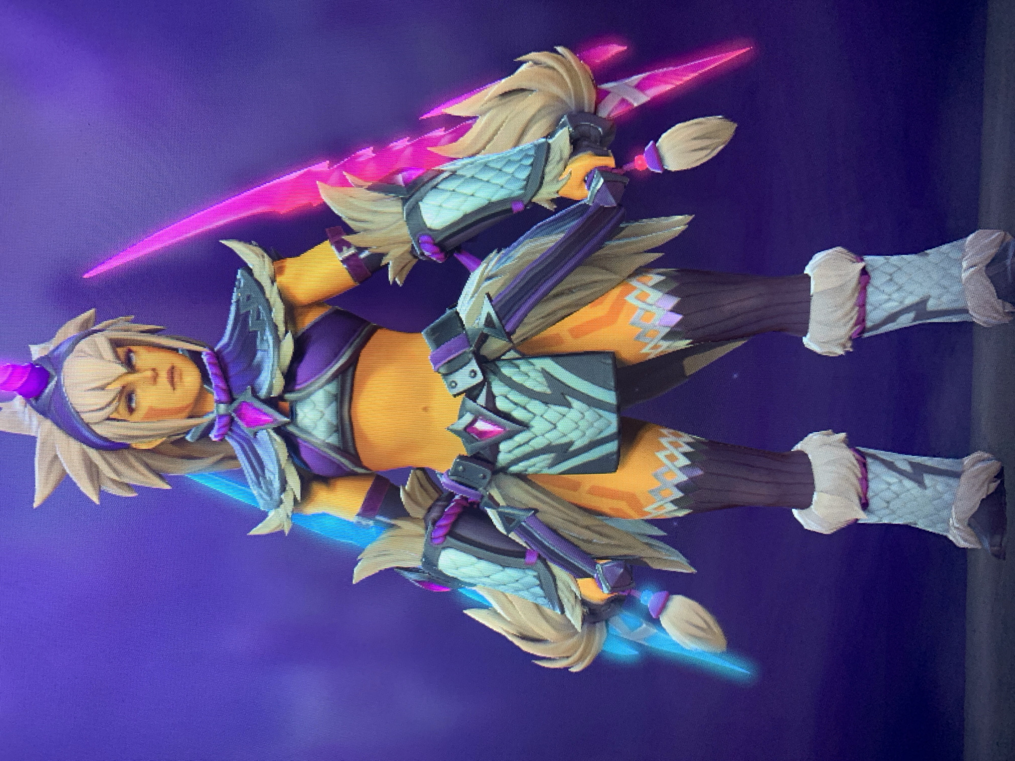 In an image of a video game character, a female warrior stands against a dark purple background. She has tanned skin, blonde hair styled in a high ponytail with bangs, and blue eyes. She wears a purple and teal outfit that includes a crop top, a skirt with a diamond pattern, and thigh-high socks. Her arms are adorned with teal and purple bracers, and she has fur-lined boots. The character wields two glowing swords, one pink and one blue, which extend from her hands. The image is lit with vibrant colors, highlighting the character's armor and weapons. The scene appears to be a character selection screen or a promotional image from a game.