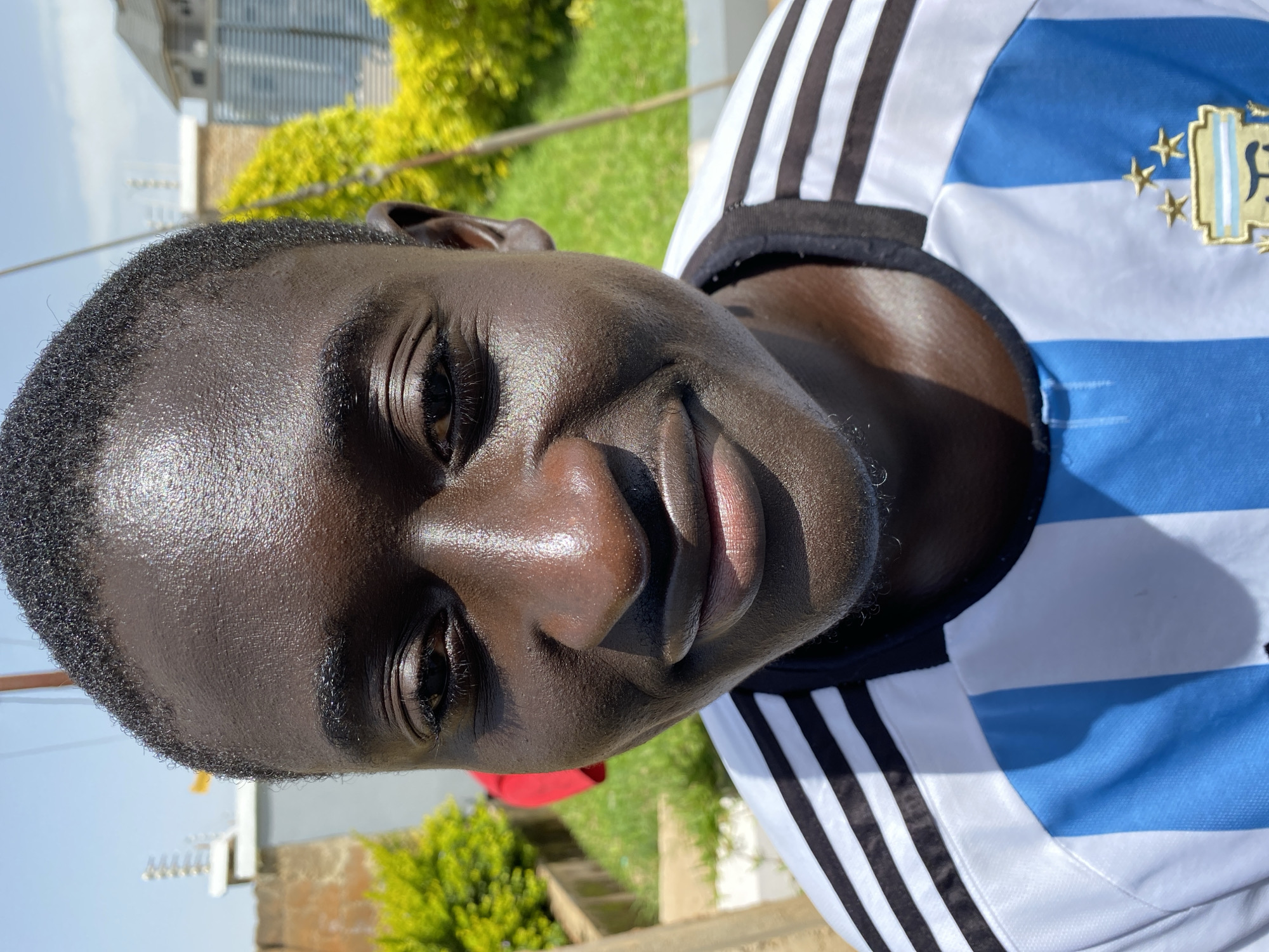 The media file shows a close-up, portrait-style image of a young Black male. He is wearing a blue and white striped jersey with black accents, which appears to be a sports jersey. The jersey has a crest with stars and some text on the right side of the chest. The subject has short, dark hair and his skin is glistening, possibly from sweat or oil, indicating a warm environment or recent activity. His eyes are dark and he has a gentle smile on his face. The background is blurred, but suggests an outdoor setting with green grass, some yellow foliage, and buildings in the distance. The lighting suggests it is daytime, likely sunny, due to the strong highlights on the subject's face. The image is taken from a low angle, looking up at the subject. The overall mood appears casual and positive. The provided location context is Bukuru, Nigeria.