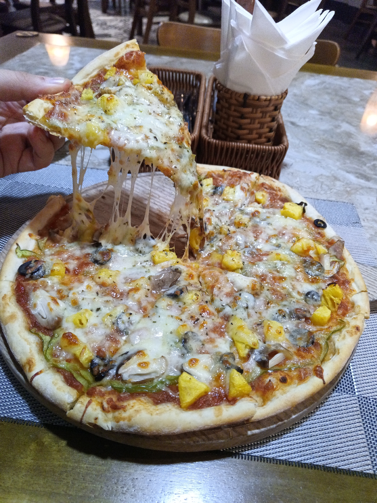 A close-up, eye-level shot shows a hand lifting a slice of pizza from a whole pizza on a wooden board. The pizza is topped with melted cheese, pineapple chunks, mushrooms, olives, and possibly chicken or seafood. The cheese stretches between the lifted slice and the rest of the pizza, indicating its gooey texture. The pizza rests on a placemat with a striped pattern. In the background, a woven basket holding napkins and a blurred restaurant interior are visible. The lighting suggests an indoor setting, possibly a restaurant in Nha Trang Ward, Vietnam.