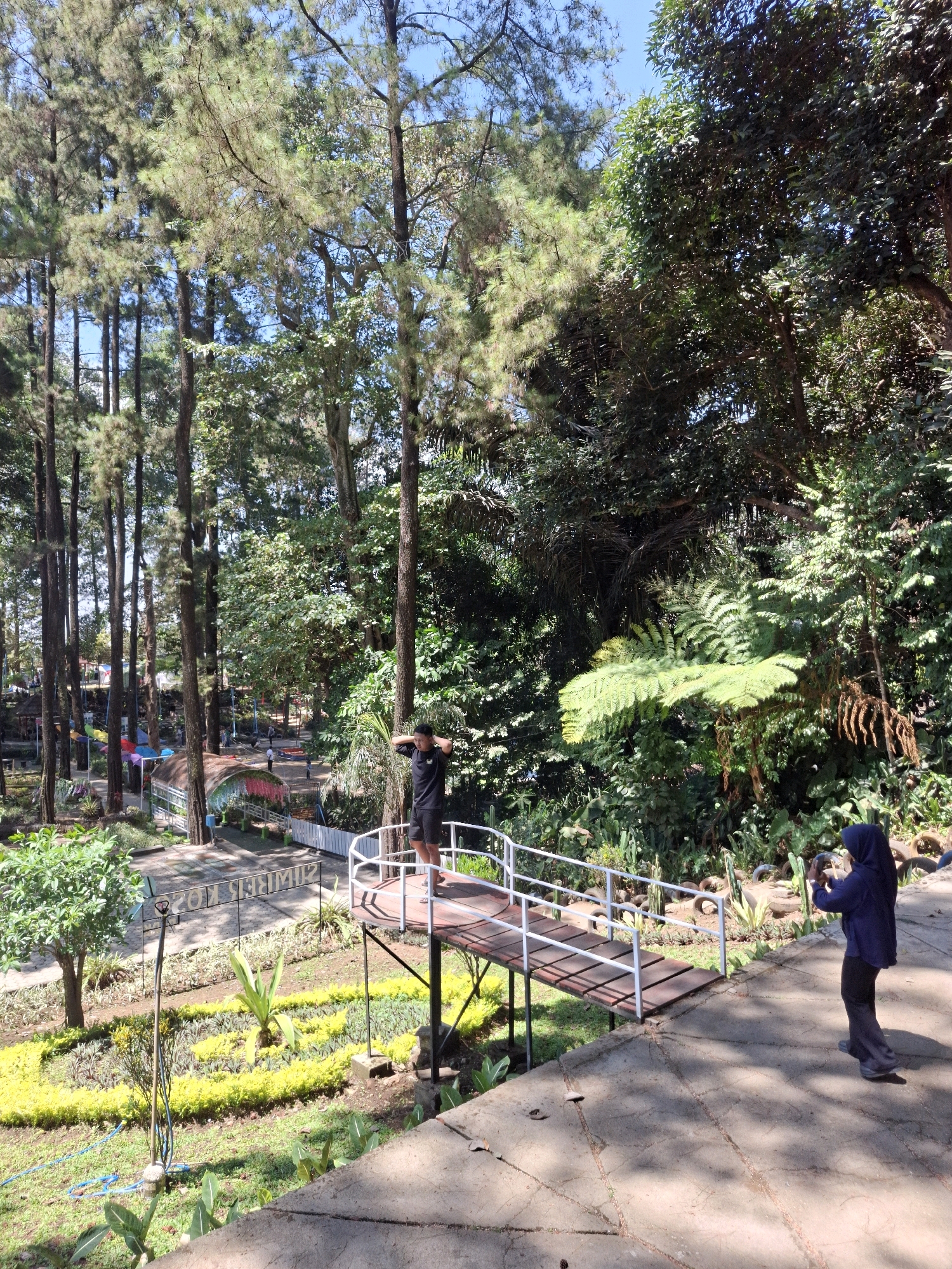 The image depicts a scenic outdoor location, possibly a park or recreational area, with lush greenery and tall trees. In the foreground, a person in a black t-shirt and shorts stands on a wooden bridge, with their hands behind their head. To the right, another person, wearing a dark blue hijab and clothing, is taking a photo with a camera. The background shows a landscaped garden with colorful flowers and plants, and further back, more trees and what appears to be a playground area with colorful umbrellas. The overall setting suggests a bright, sunny day. The location is identified as Girikerto, Indonesia.