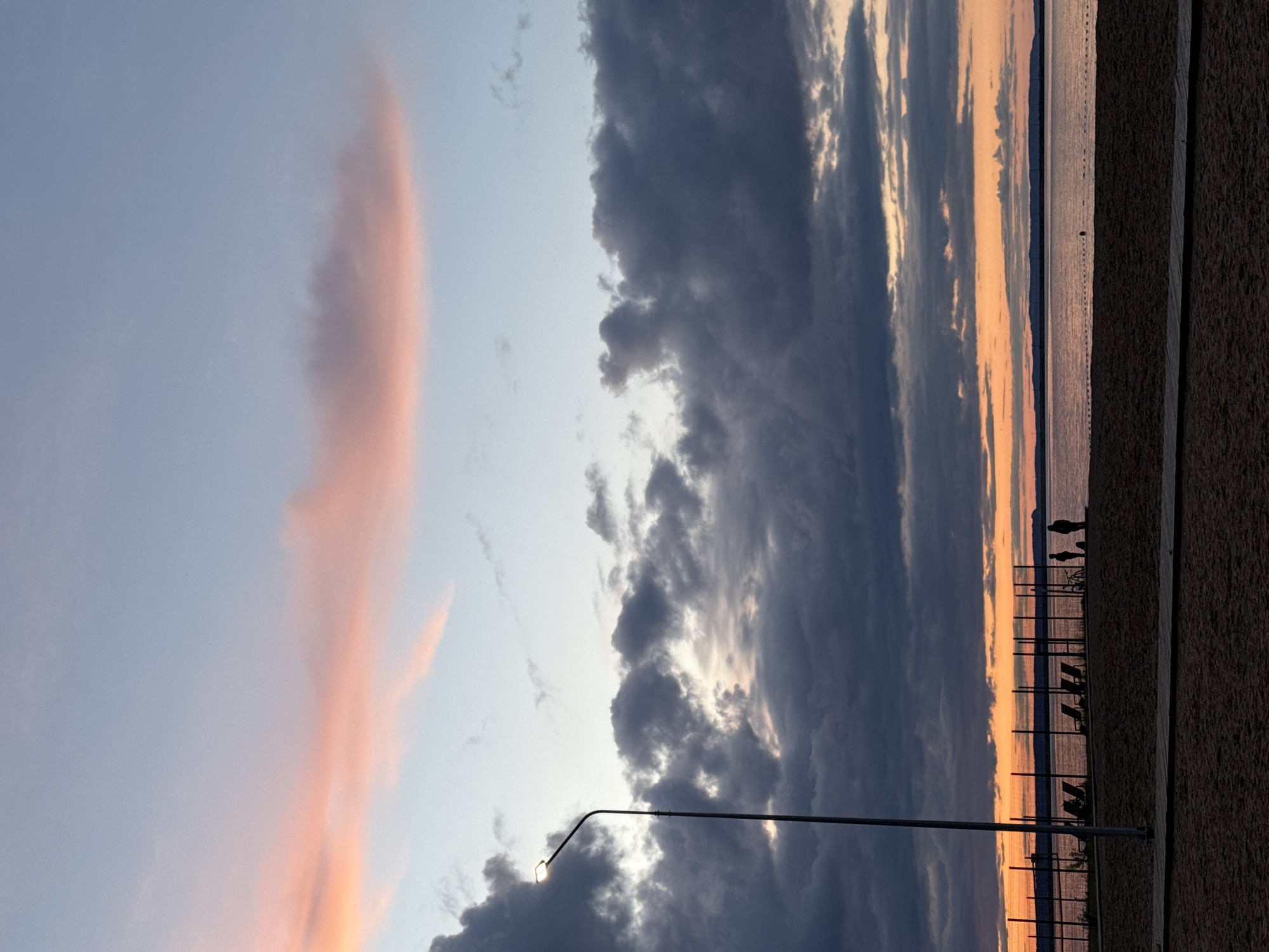 The image depicts a sunset scene in what appears to be the city of Chaikovsky, Russia, judging by geographical details.
The sky is partially cloudy with streaks of pink and orange hues indicating the sunset. To the right is a body of water reflecting the sunset colors.
A street lamp is visible in the foreground, positioned at an angle. A fence runs along the waterfront. Silhouetted figures of people are discernible along the fence.
