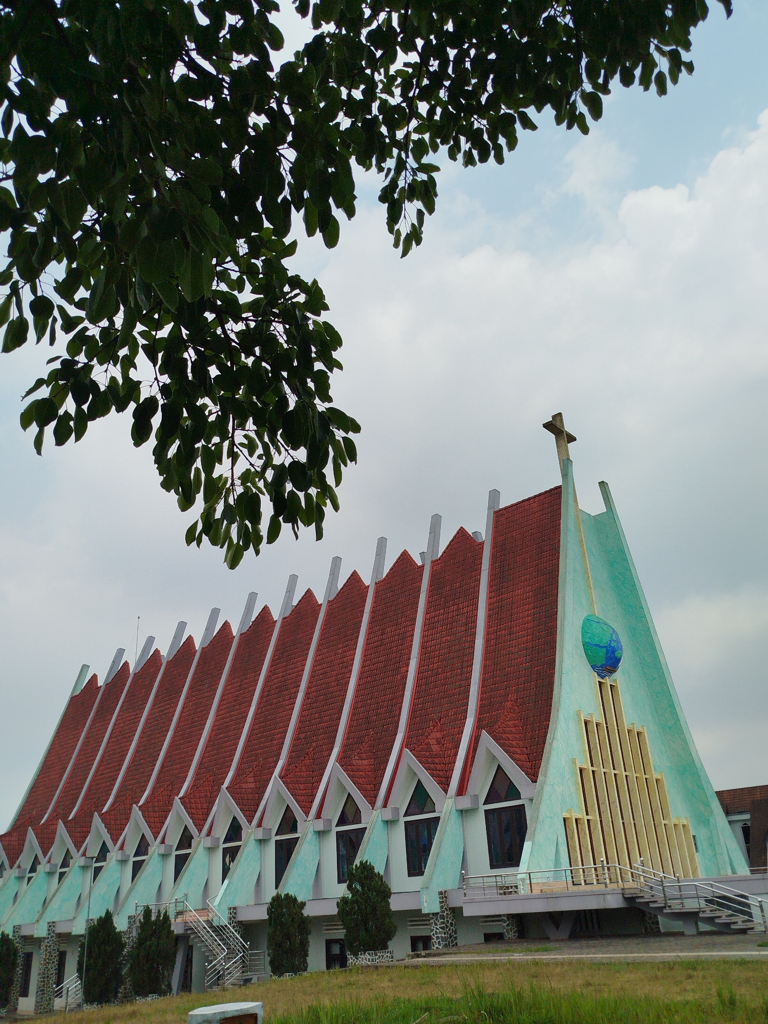 The image shows a distinctive church building in Lang Biang - Đà Lạt, Vietnam. The church has a red tiled roof with a series of triangular peaks separated by white accents. The exterior walls are light blue. At the front of the church is a tall section with a cross at the top. A blue globe is mounted on the front, beneath which are vertical beige stripes. The church is situated on a grassy slope with trees. A tree branch with green leaves occupies the upper left corner of the image. The sky is overcast.