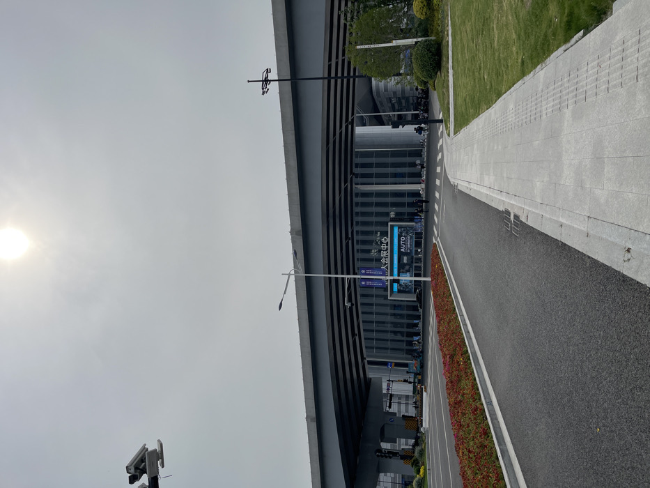 The image shows a street in Hang Zhou Shi, China, located at coordinates 30.273 latitude, 120.419 longitude. The scene is under a bright, sunny sky with no clouds. A large, modern, glass-fronted building stands in the middle of the road. The building has a sign that reads "AUTO" and Chinese text, indicating it is a car dealership. The street is asphalt and has a pedestrian walkway with a sidewalk on the right. Several people are walking in the street and several cars are parked in the distance. The street has a few streetlights and a large, concrete overpass. Trees and green grass are on the right and a bed of red flowers borders the left side of the street. There is a security camera on a pole on the left side of the image.