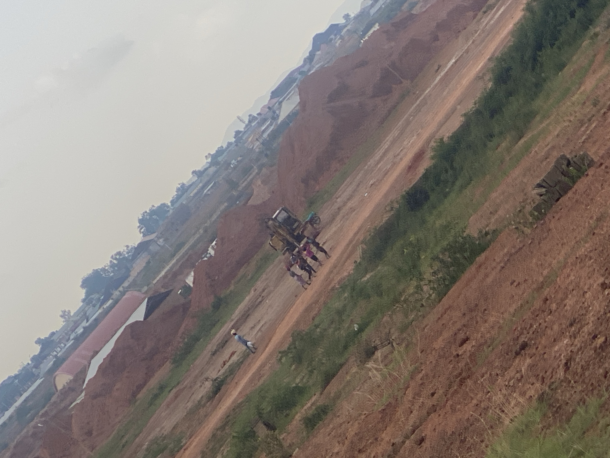 The image depicts a construction or excavation site in the city of Jos, Nigeria. The terrain is characterized by large mounds of reddish-brown earth, indicative of soil disturbance. Several individuals are present at the scene, engaged in what appears to be work or activity related to the site. Some are gathered around a piece of heavy machinery, possibly an excavator. The environment suggests an ongoing development or land modification project, with scattered vegetation and distant buildings visible in the background. The sky is overcast, contributing to a hazy atmosphere.