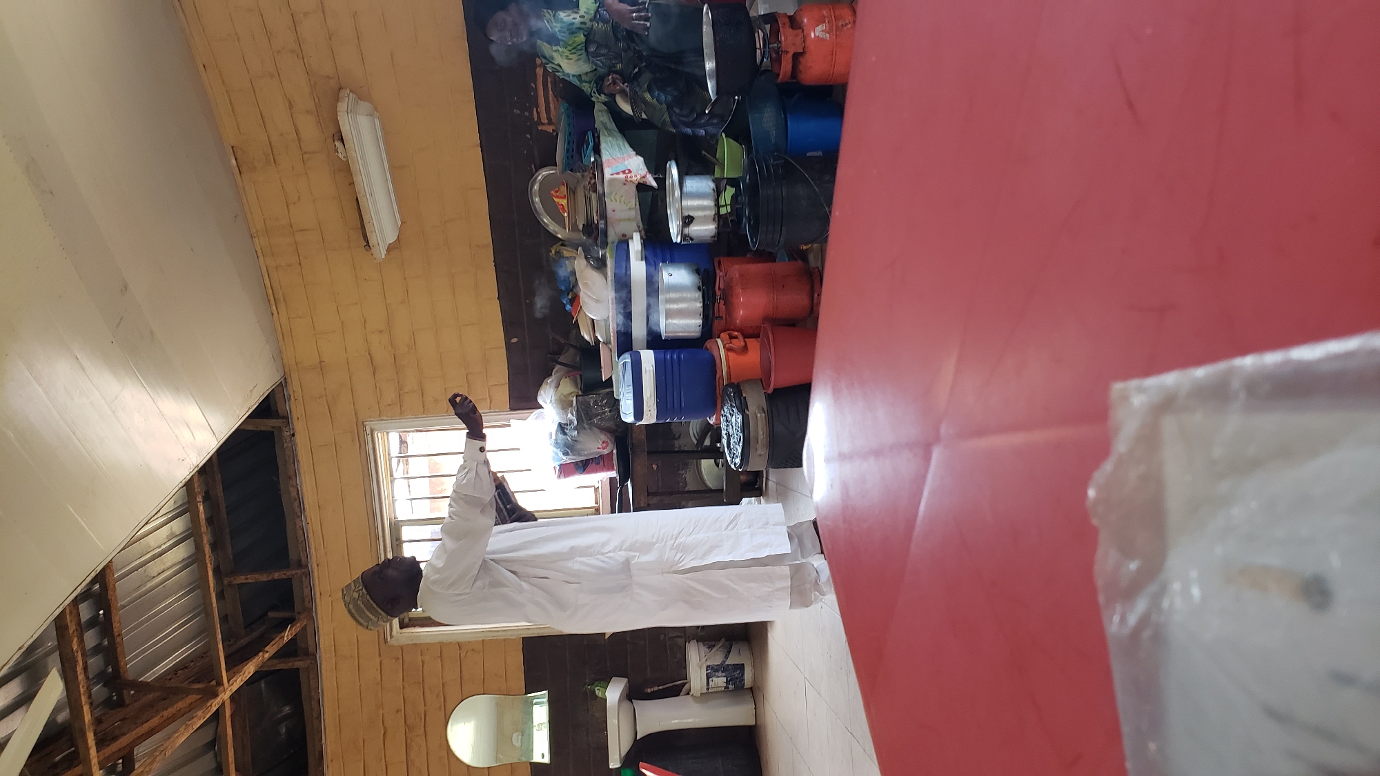 The image depicts a man wearing a white robe and a patterned cap, standing with his arms raised as if in prayer or supplication. He is positioned in what appears to be an indoor space, possibly a workshop or storage area, given the collection of items behind him. The background is filled with various objects, including stacked gas cylinders, pots, coolers, and other containers. A red table is prominently visible in the foreground, partially obscuring the scene. The walls are made of yellow brick, and a window with horizontal blinds is visible on the right. Below the window, a toilet and a bucket are situated on the floor. The overall impression is of a utilitarian or storage space, possibly in a community setting. The man's posture suggests a moment of devotion or deep thought. The lighting appears to be from artificial sources, though a hint of natural light might be coming from the window. No text is discernible in the image. The image is oriented sideways, with the man facing towards the left of the frame.
