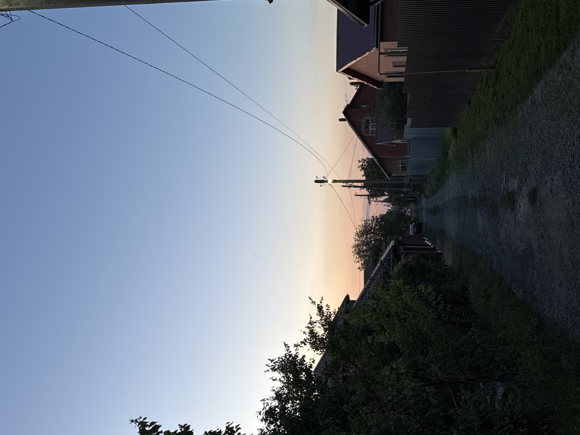 The image depicts a rural street in Grushevskaya, Russia, at dusk. The sky transitions from a pale blue at the top to a soft gradient of pink and orange near the horizon. A gravel road runs through the center of the frame, flanked by houses and fences on either side. To the left, dense green foliage obscures some of the houses. The houses on the right are more visible, featuring brick exteriors and dark-colored roofs. Overhead, power lines are strung between utility poles. A single car is visible in the distance down the road. The overall atmosphere is quiet and serene, characteristic of a rural setting during twilight.