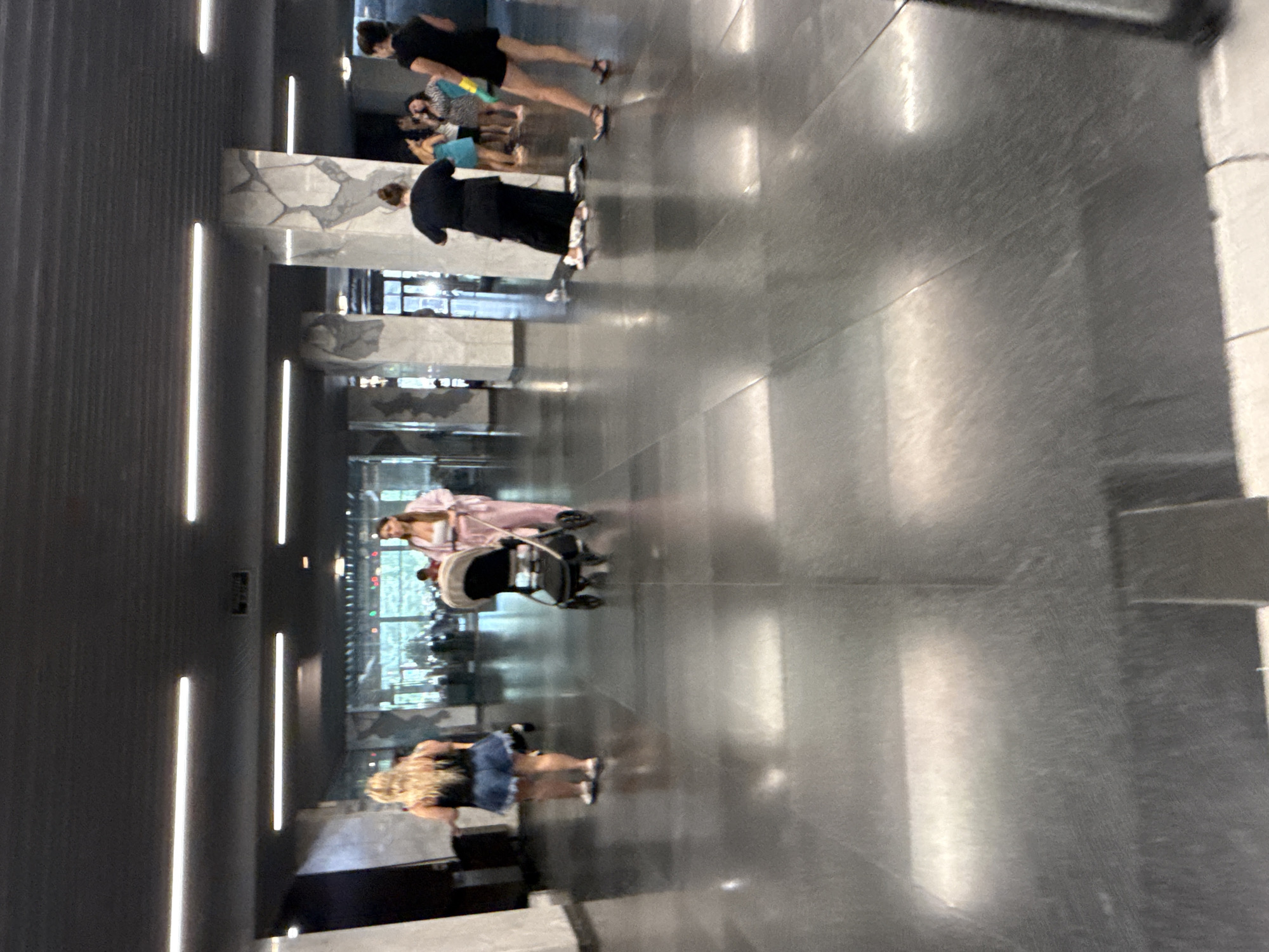 In a spacious, modern interior with a polished dark floor and a high, dark ceiling featuring recessed linear lighting, a group of people are visible. On the left, a woman with blonde hair is seen from the back, wearing a denim skirt and a dark top, standing. Further into the hallway, a woman in a pink loungewear set pushes a beige and black stroller. To the right, a person in a long black dress is partially visible, with another individual in a black top and shorts and sandals to their right. Several other figures are dispersed in the background, some appearing to be standing and conversing. The architecture includes large, light-colored columns with abstract patterns. The overall environment suggests a public indoor space, possibly a lobby or a wide corridor. There are no identifiable landmarks that would specifically place this scene in Yalta, Ukraine.