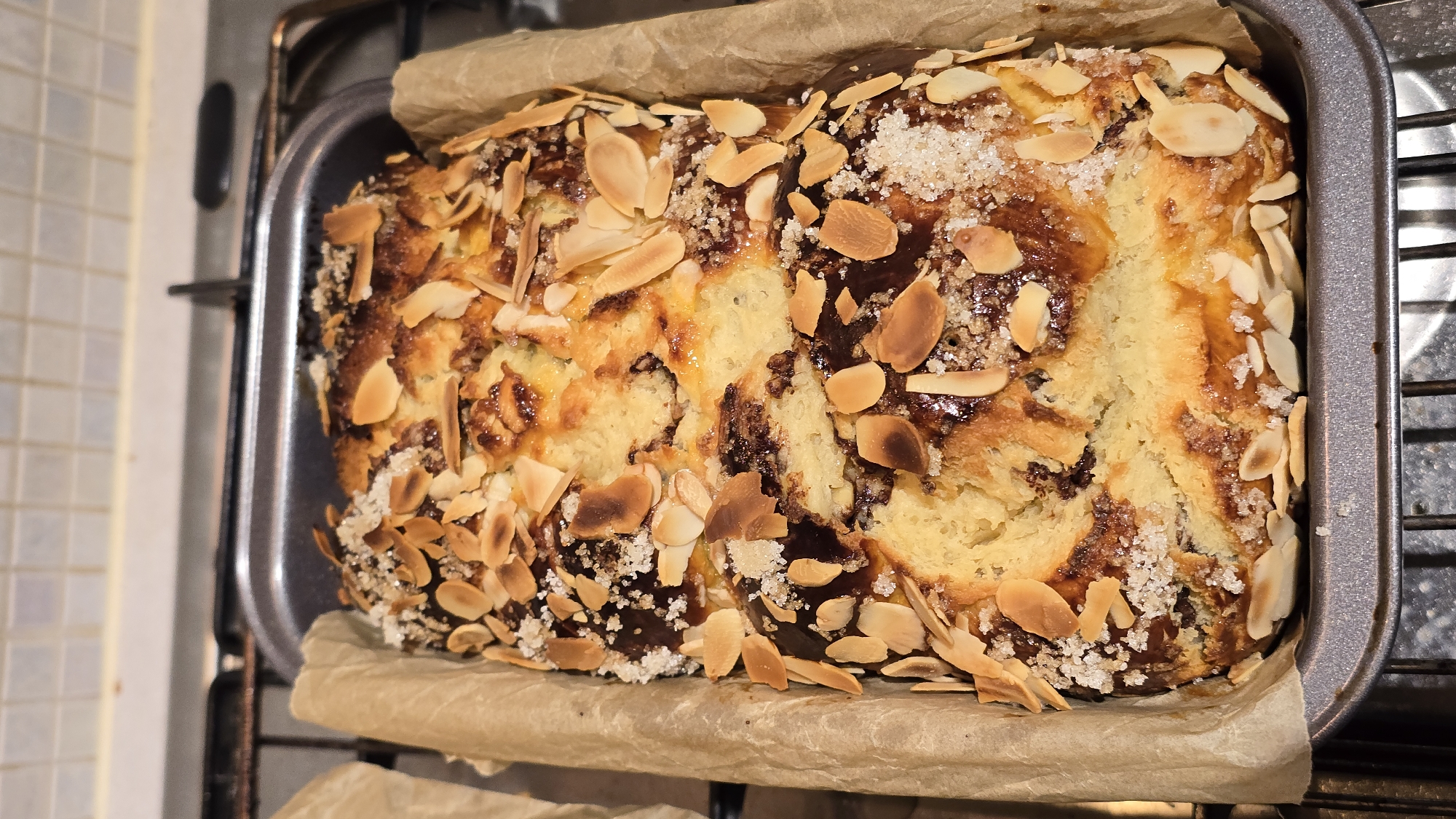 A freshly baked sweet bread loaf is presented in a metal baking pan lined with parchment paper. The loaf, with a golden-brown crust speckled with toasted almond slices and coarse sugar crystals, sits atop a stovetop range. The kitchen, likely in a residential setting, is uncluttered.