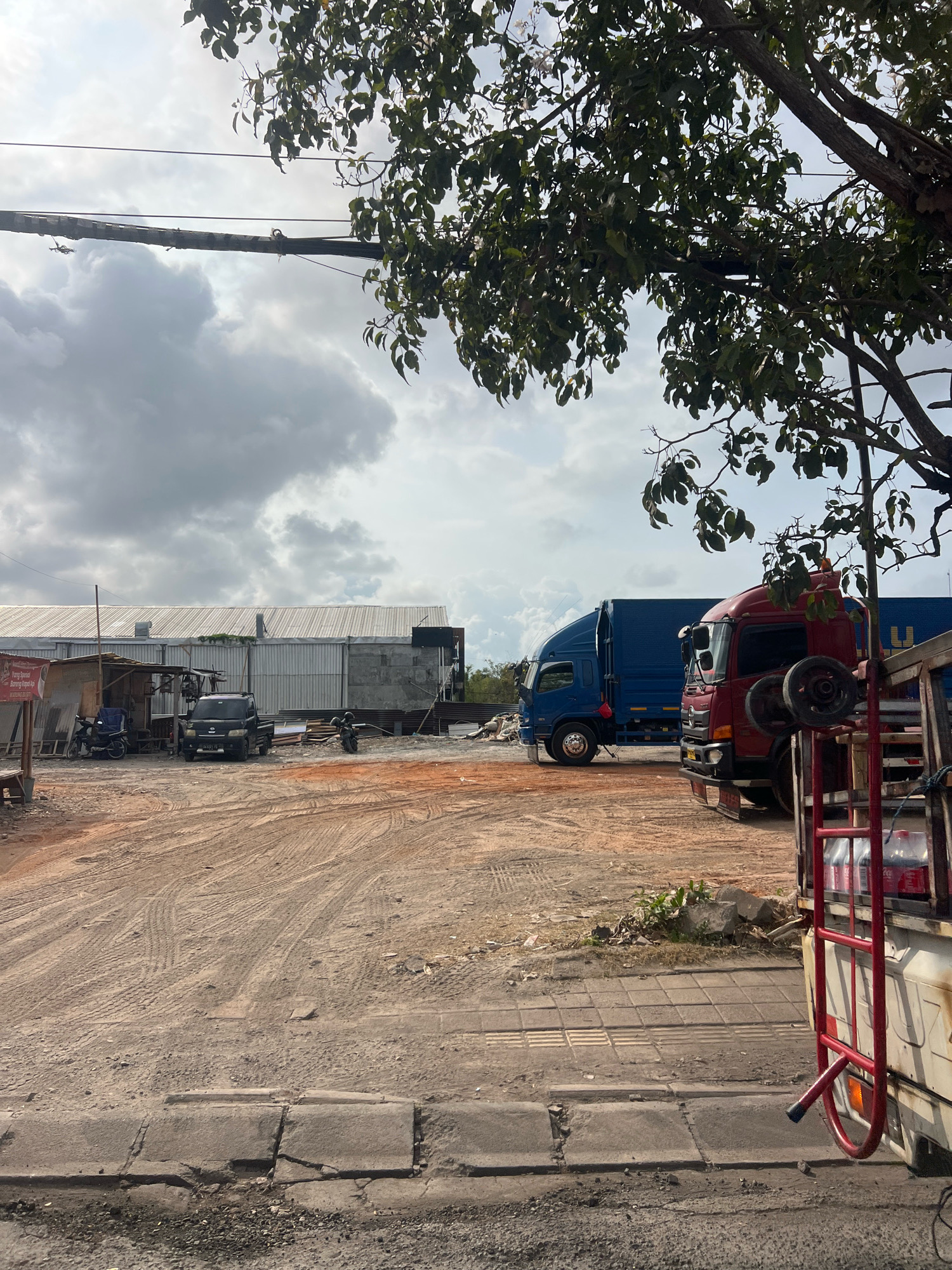 The image captures an outdoor scene on a cloudy day, likely in Denpasar, Indonesia, given the context provided. The foreground is dominated by a dusty, uneven ground surface with tire tracks and scattered debris. A portion of a white vehicle with a red ladder rack is visible on the right side, carrying two bottles of Coca-Cola. In the mid-ground, a red truck and a blue truck are parked on the dusty terrain. Behind the trucks, a large, long building with a corrugated metal roof is visible, appearing to be a warehouse or industrial structure. To the left of the building, a small, makeshift shelter is present, with a dark-colored utility vehicle and a motorcycle parked nearby. Scattered construction materials, including metal sheeting and lumber, are visible throughout the scene. The sky is overcast with dramatic, grey clouds.