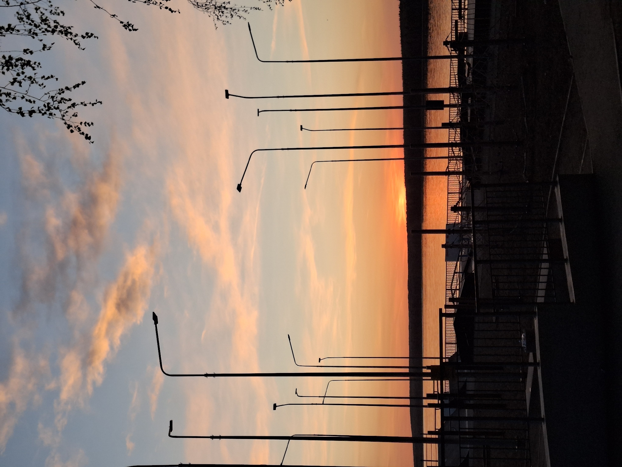 The image depicts a sunset over the Kama River in Chaikovsky, Russia. In the foreground, there's a row of streetlights with a branch of a tree at the upper left. Silhouetted fencing, staircases, and architectural elements are visible to the right. The background features the Kama River reflecting a vibrant orange sunset, with the opposite shoreline visible as a dark band. The sky above displays pastel shades of blue, pink, and orange, interspersed with clouds.