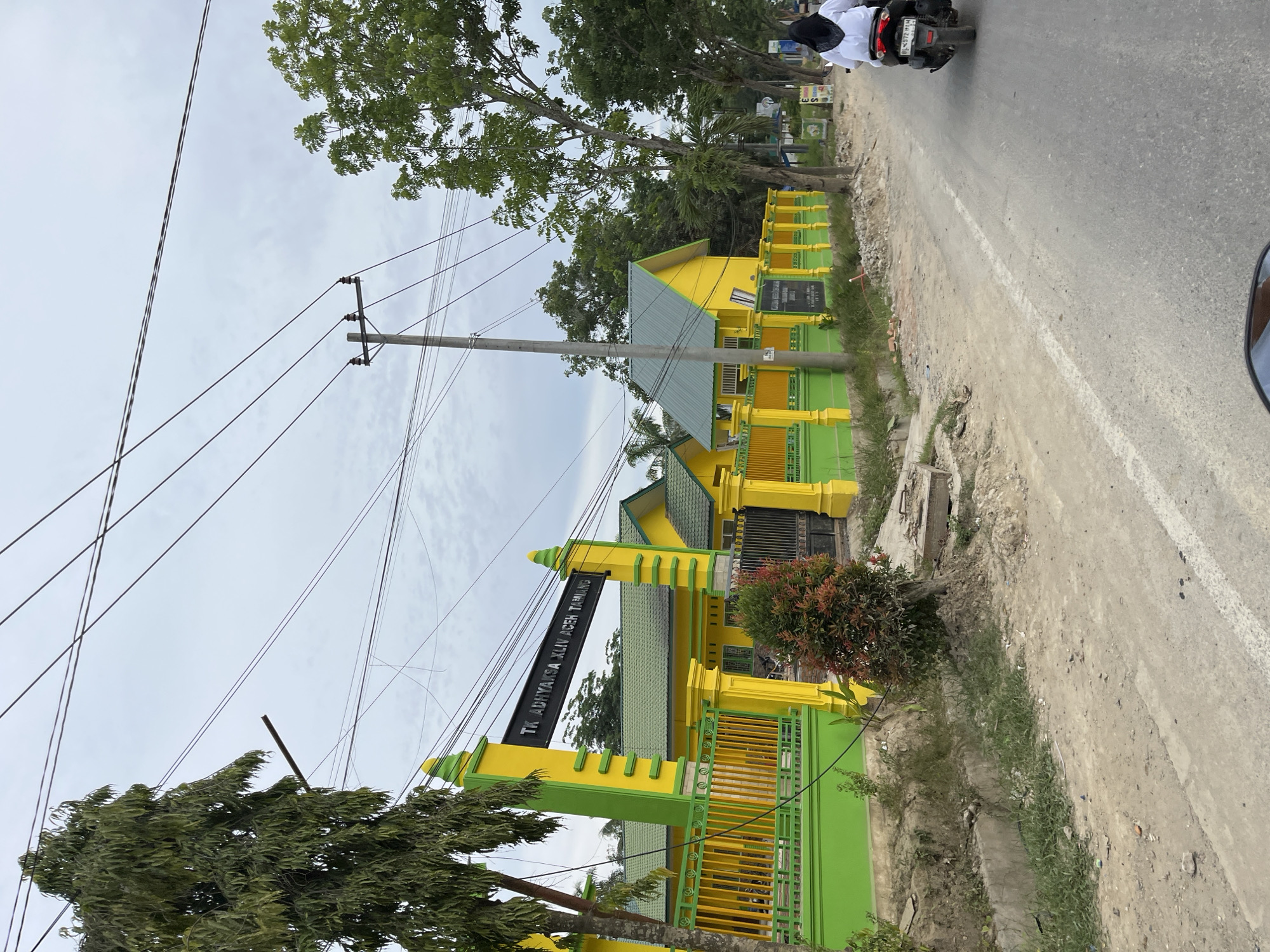 In Bundar, Indonesia, a school building with bright yellow walls and green roofs is visible, bordered by a green and yellow fence. The building has a sign that reads "TK ADHYAKSA XLIV ACEH TAMIANG." A person wearing a white hijab and top is riding a motorcycle away from the camera on a paved road in front of the school. The sky is overcast, and power lines are visible overhead. The surrounding area appears to be a mix of trees and open land.