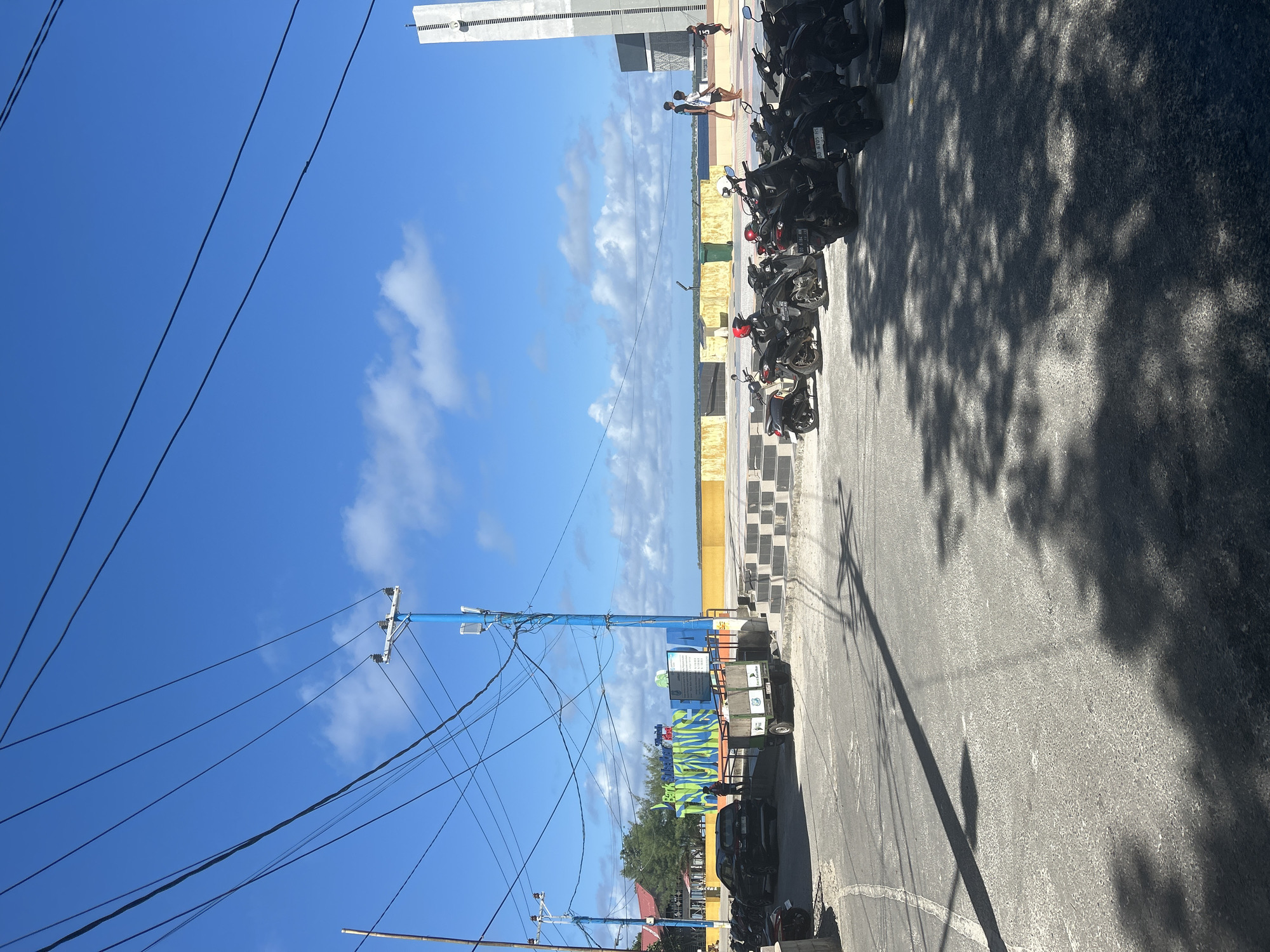 In Bira, Indonesia, a paved area is filled with parked motorcycles under a clear blue sky with scattered clouds. A blue utility pole with numerous wires stands prominently, casting shadows on the ground. In the background, a yellow wall borders the sea, and a modern, tall structure, possibly a monument or part of a building, is visible on the right. Several people are walking near the water's edge. To the left, a black car is parked near a sign with colorful lettering, and a small utility trailer is situated nearby. The scene depicts a bustling outdoor area with elements of urban infrastructure and coastal landscape.