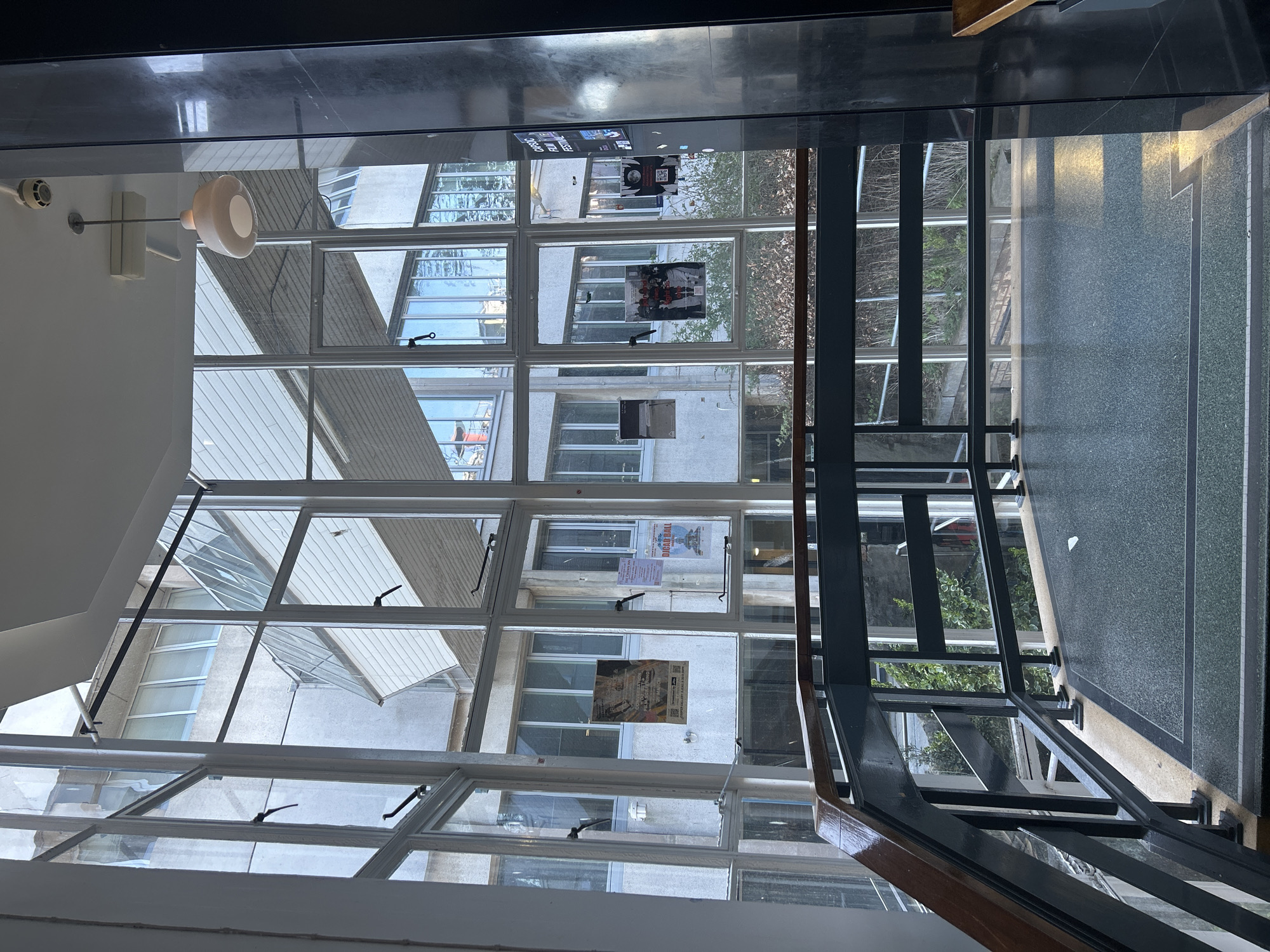 The image, oriented as a landscape but appearing to be a rotated portrait view, captures an indoor architectural space looking out through expansive windows. In the foreground, on the left, a light-colored interior wall features a modern, cream-colored dome light fixture suspended by a rod, and a white smoke detector or fire alarm. Above, a dark, highly reflective ceiling creates a mirror-like surface. To the right, a robust, dark grey metal staircase railing with a warm brown wooden handrail descends, bordering a dark grey, speckled floor with lighter grey striping.
The majority of the frame is dominated by a tall, multi-paned window structure, comprising numerous rectangular glass sections framed in white or light-colored sashes. Through these windows, an exterior courtyard or lightwell is visible, bordered by several multi-story, light-colored buildings. These external structures feature their own arrays of windows, some of which are partially open. Small patches of green foliage, likely from trees or bushes, can be glimpsed outside.
Several posters are affixed to the exterior walls of the buildings beyond the windows. One clearly legible poster advertises a "CLASSIC FILM FESTIVAL." Another prominent poster below it features the bold text "DOG EAT DOG." A third, smaller poster, less distinct, includes what appears to be event information and a QR code. The scene is brightly lit by natural light, suggesting it is daytime, possibly with overcast skies given the soft illumination and lack of sharp shadows. No people are visible in the image.
