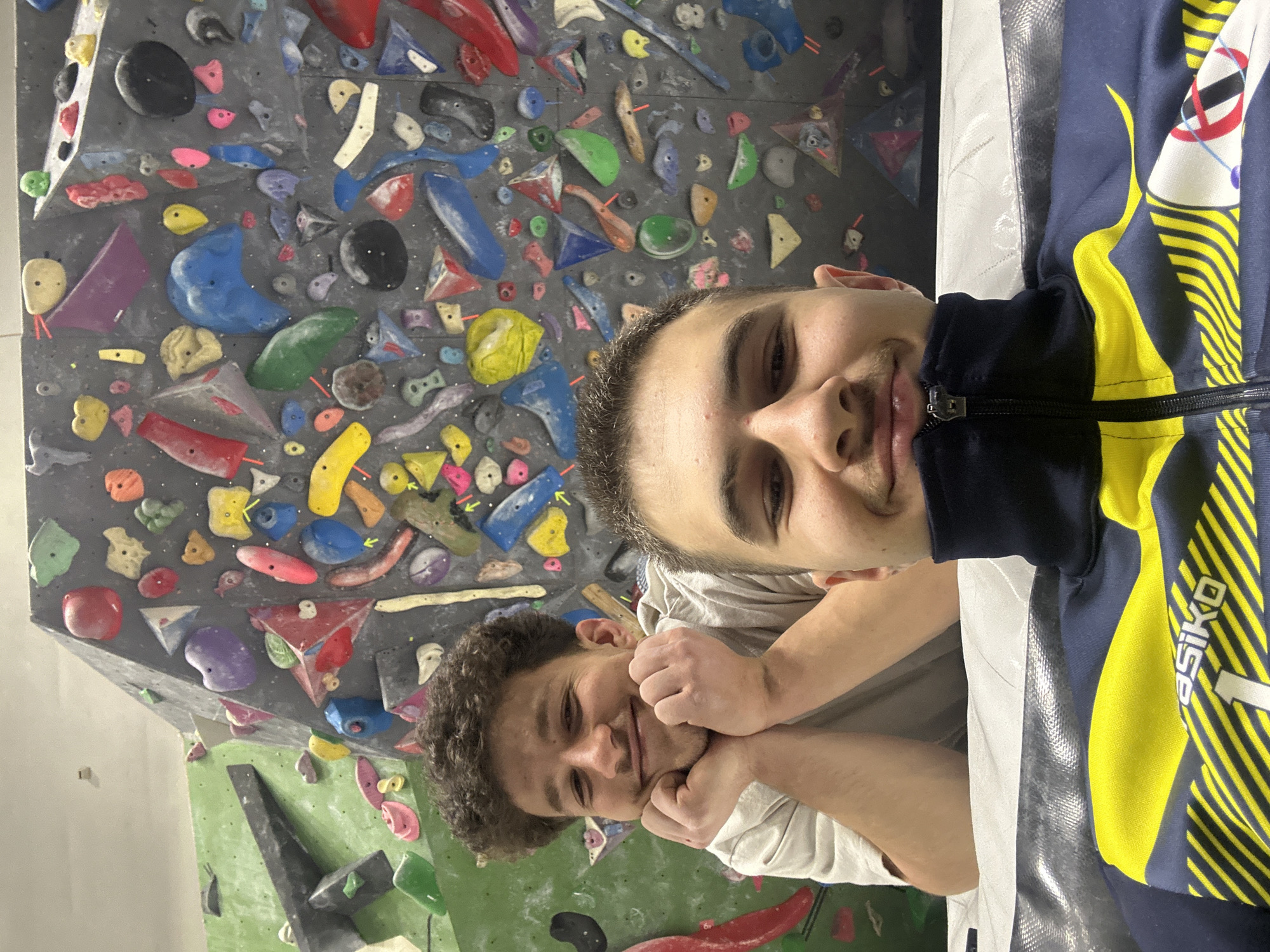A close-up photograph depicts two males within an indoor bouldering gym in Sofia, Bulgaria. In the immediate foreground, a male's face is visible from the nose down, wearing a navy blue and yellow athletic jacket with a 'siko' logo and diagonal stripes; he displays a slight smile and looks directly forward. Behind him, a second male with curly brown hair, wearing a light-colored t-shirt, smiles broadly with his hands clasped under his chin. Both individuals look directly at the camera. The background features an angled grey and green bouldering wall densely covered with numerous multi-colored climbing holds of various shapes and sizes, indicating an active climbing environment. A white and grey crash pad is partially visible beneath the individuals.