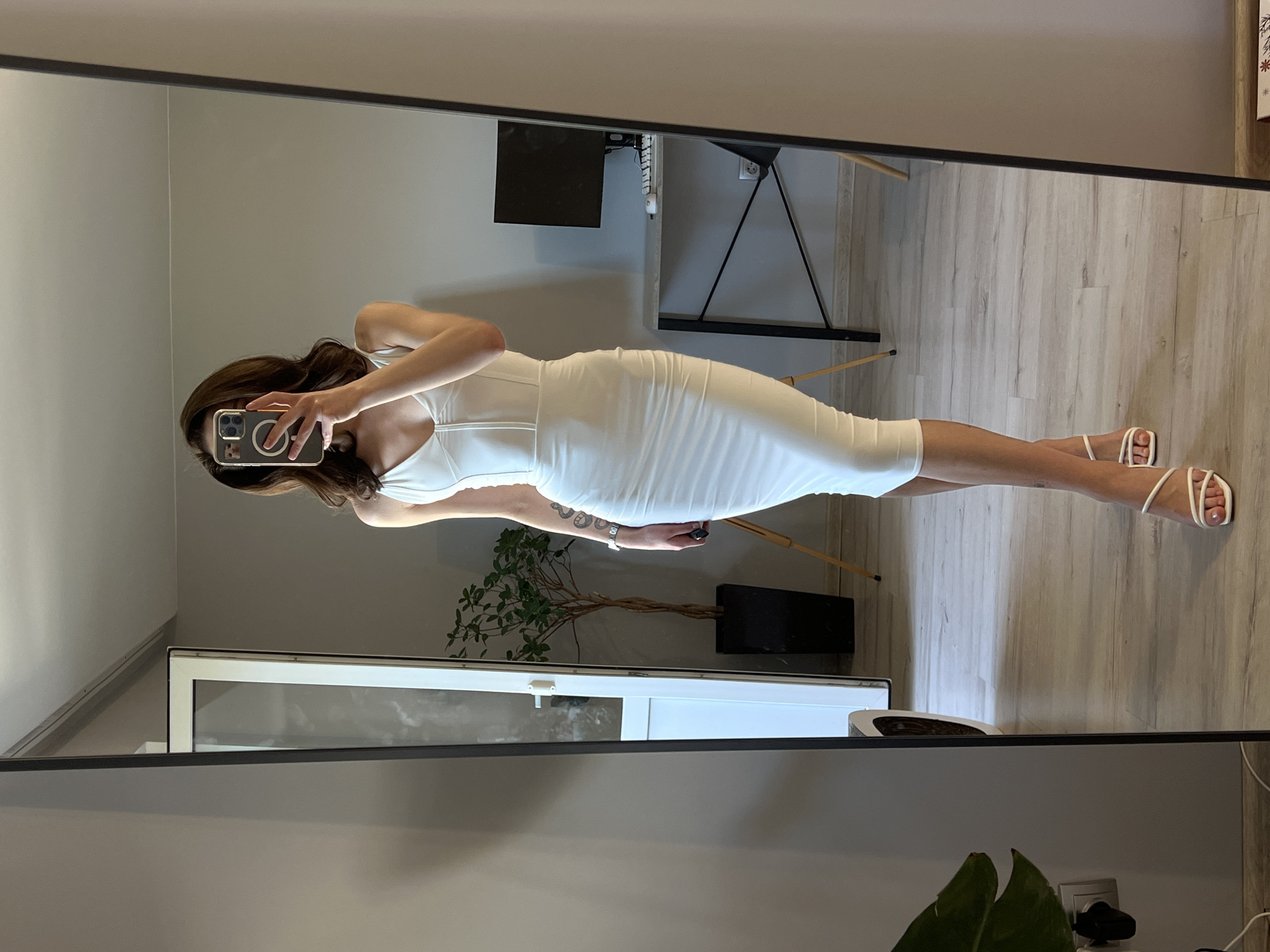 A young woman, with medium-length brown hair and her face obscured by her phone, stands in front of a full-length mirror, taking a selfie. She is wearing a tight, white, ruched, midi-length dress with a sweetheart neckline and white strappy heeled sandals. Her left arm is raised, holding the phone, and her right arm hangs loosely at her side. A tattoo is visible on her left forearm.
The background reveals a room with light grey walls and a light wood-laminate floor. To her left, a large mirror reflects the scene. Beyond the mirror, a glass door leads to an outdoor area, and a tall potted plant with green leaves stands beside it. To her right, a modern desk with a black computer monitor, keyboard, and mouse is visible, along with a black office chair. The overall impression is that of a domestic setting, likely a bedroom or dressing room. This scene is not specifically identifiable as being in the city of Ternopil, Ukraine, but rather a general interior setting.